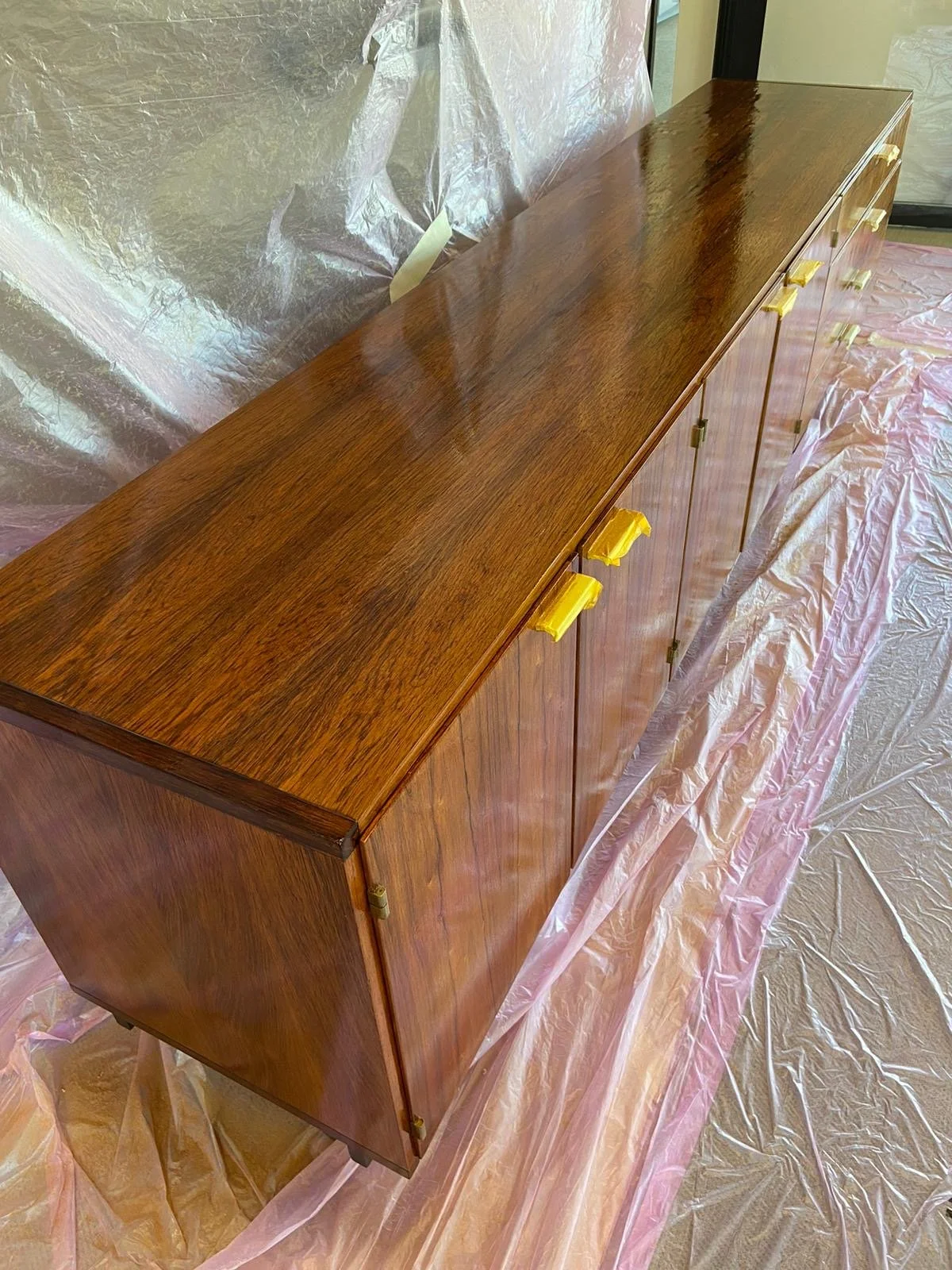A vintage wooden sideboard with brass hinges and yellow-painted handle knobs, covered with a protective plastic sheet, in a room with pink plastic floor covering.
