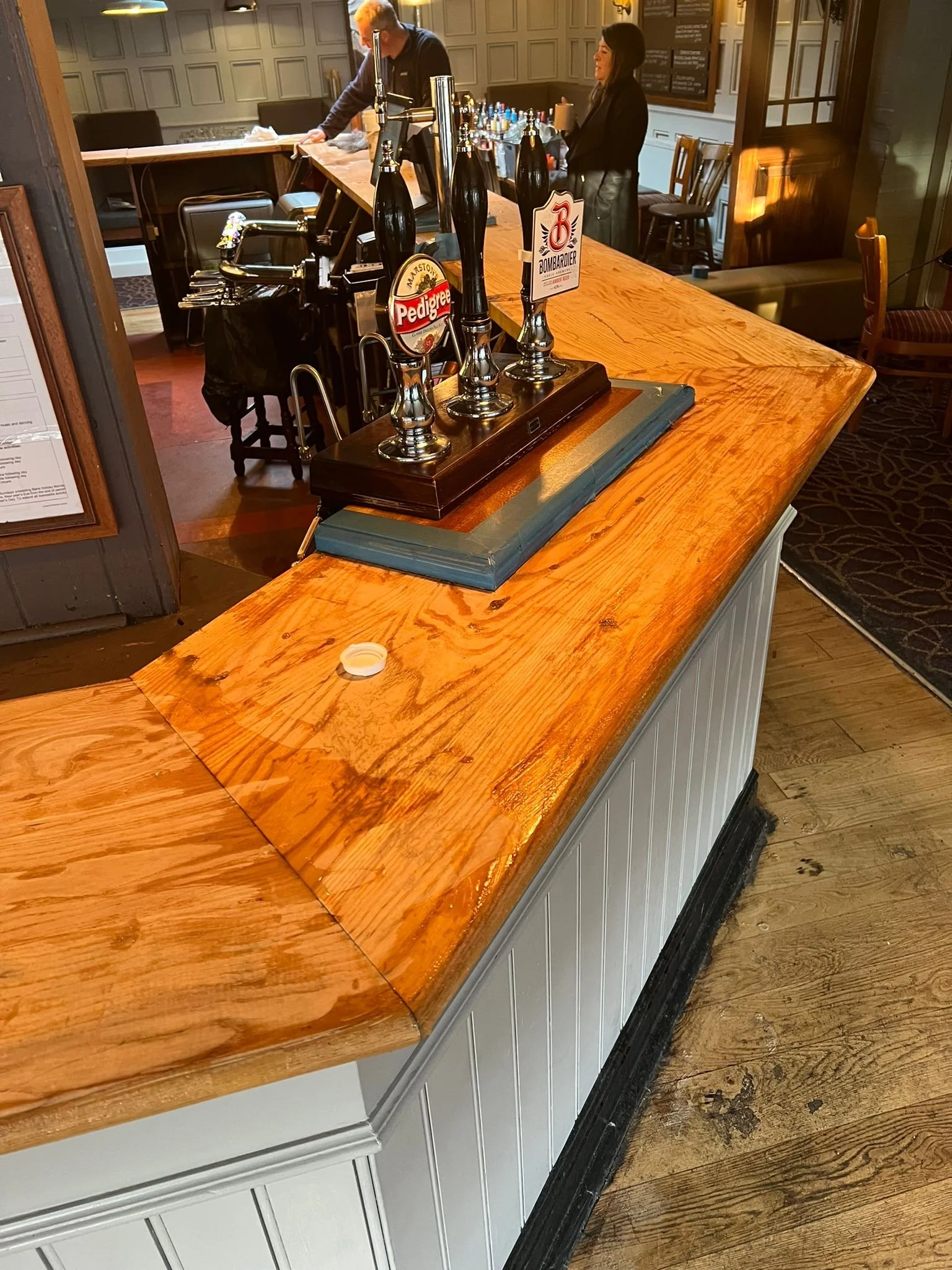 Empty wooden bar counter with beer taps, with two patrons in the background. One person is pouring a drink and another is holding a cup, inside a cozy pub with paneled walls and wooden furniture.