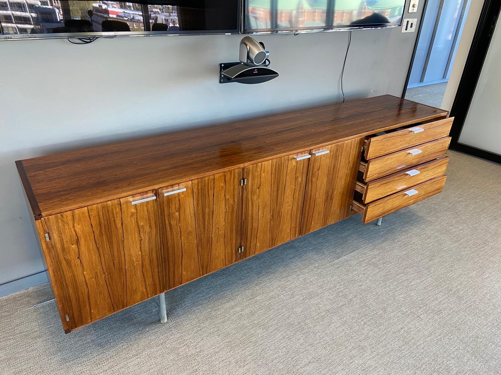 A mid-century modern wooden sideboard with multiple doors and three open drawers, topped with a flat wooden surface, located in an office or conference room with a wall-mounted TV, speaker, and power outlets in the background.