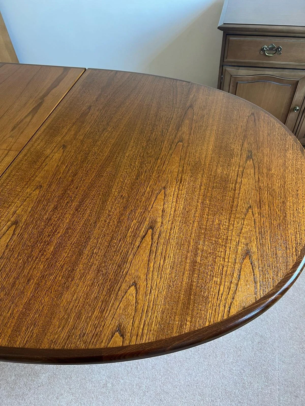 Close-up of a wooden dining table with a natural finish and visible wood grain in a room with beige carpet and a matching wooden cabinet in the background.
