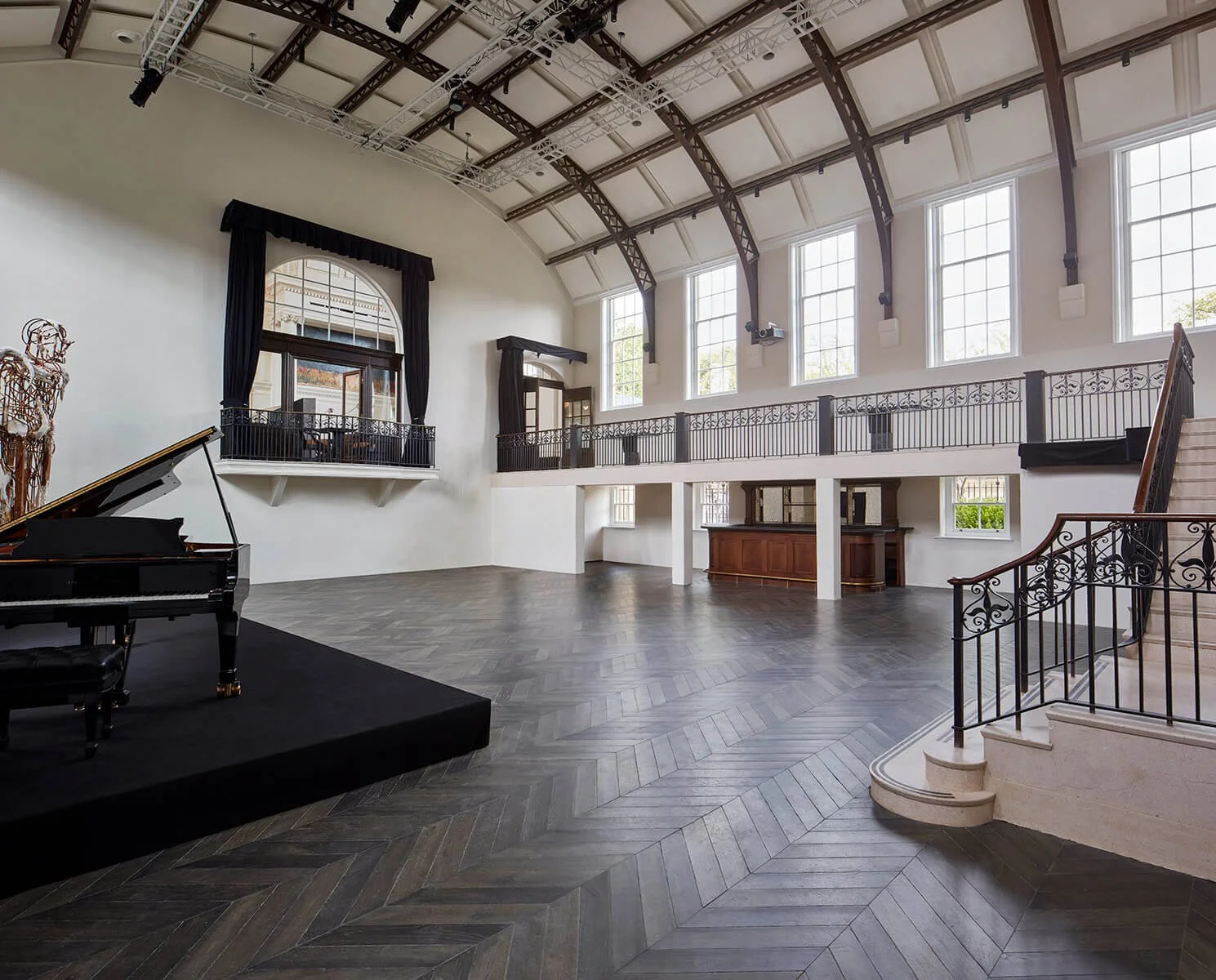 Large, elegant indoor space with high arched ceiling, large windows, hardwood flooring, grand piano on a black platform, and a staircase with decorative railing.