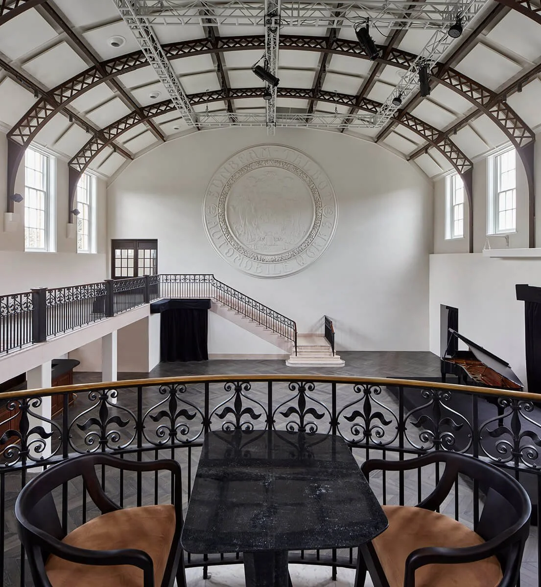 Interior of a large hall with high arched ceiling, large windows on sides, a staircase, a grand piano, and a decorative wall medallion.