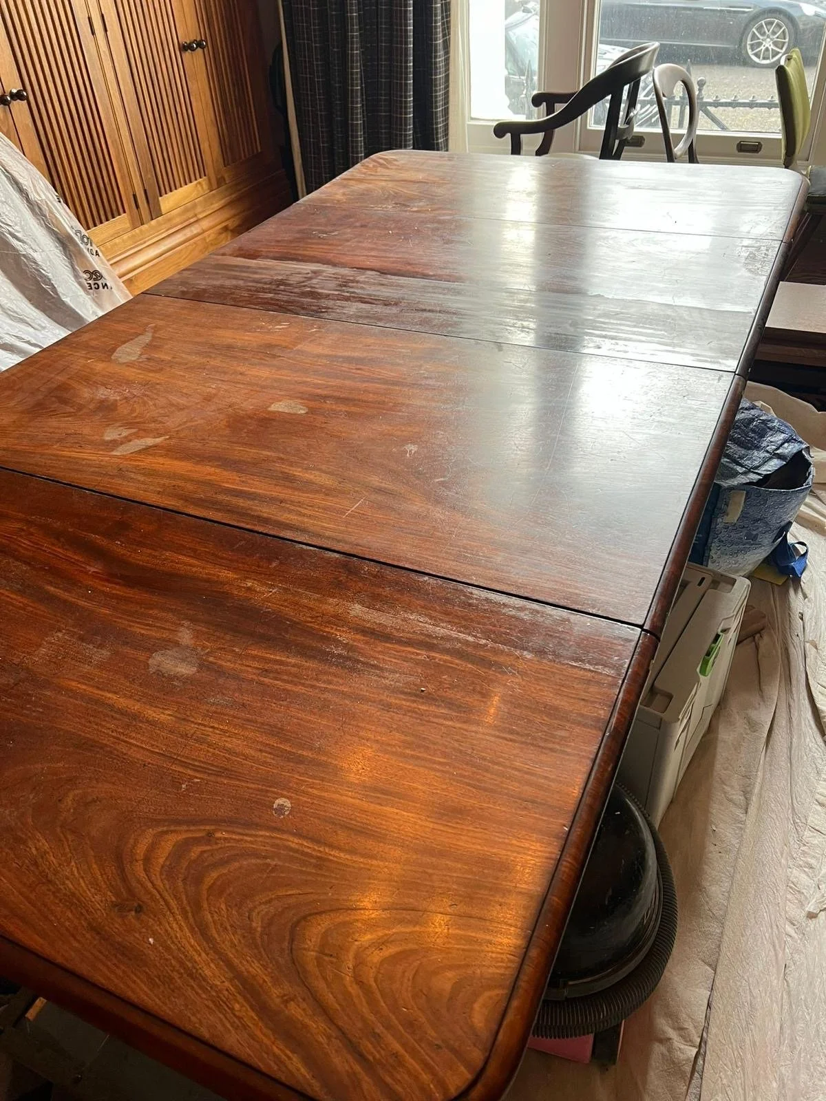 A wooden dining table with a polished surface, showing some scratches and marks, situated in a room with a window and various chairs around it.