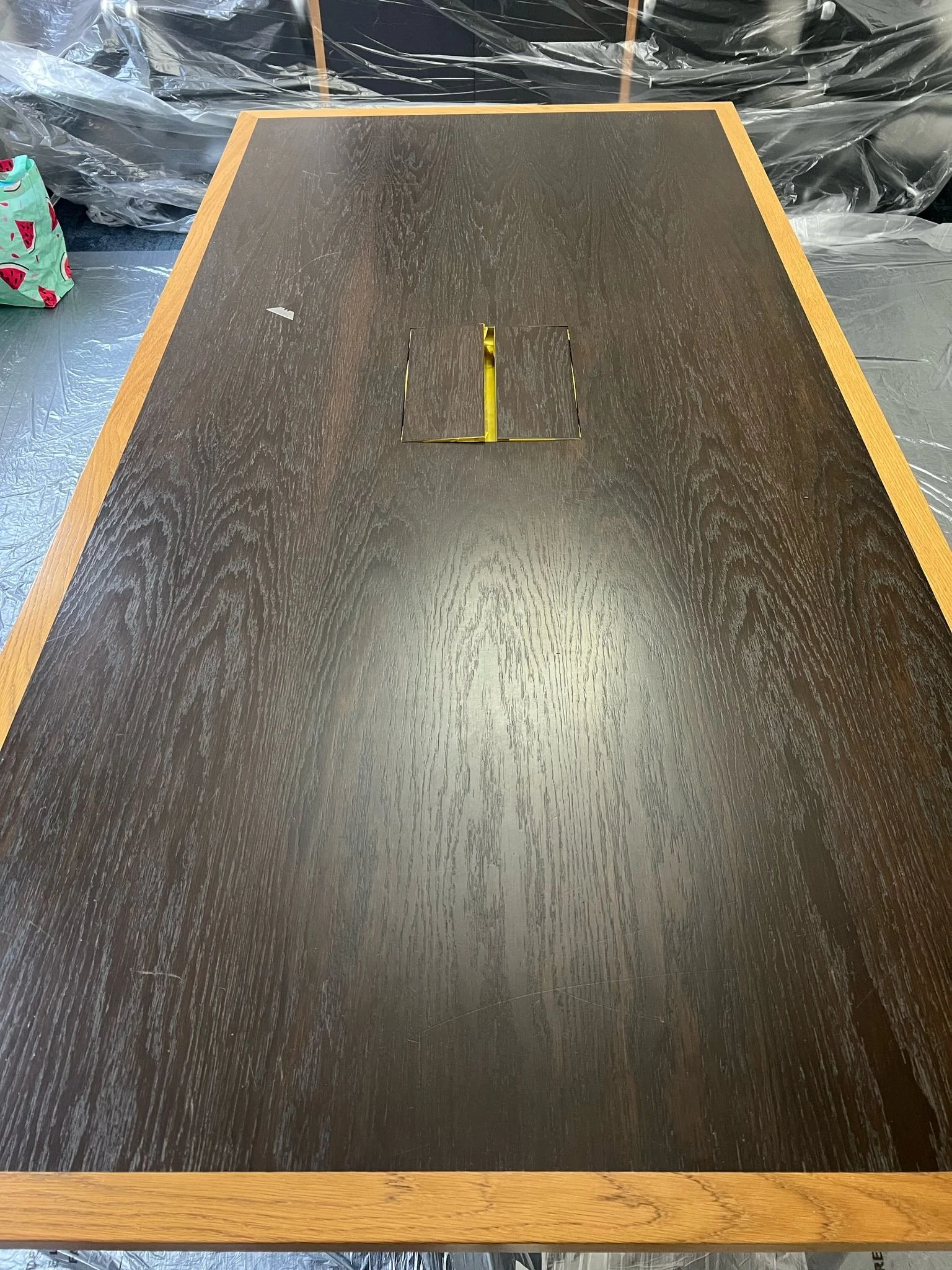 A dark wood conference table with a rectangular inlay in the center, surrounded by a light wood border, in a room with plastic-covered walls or partitions.