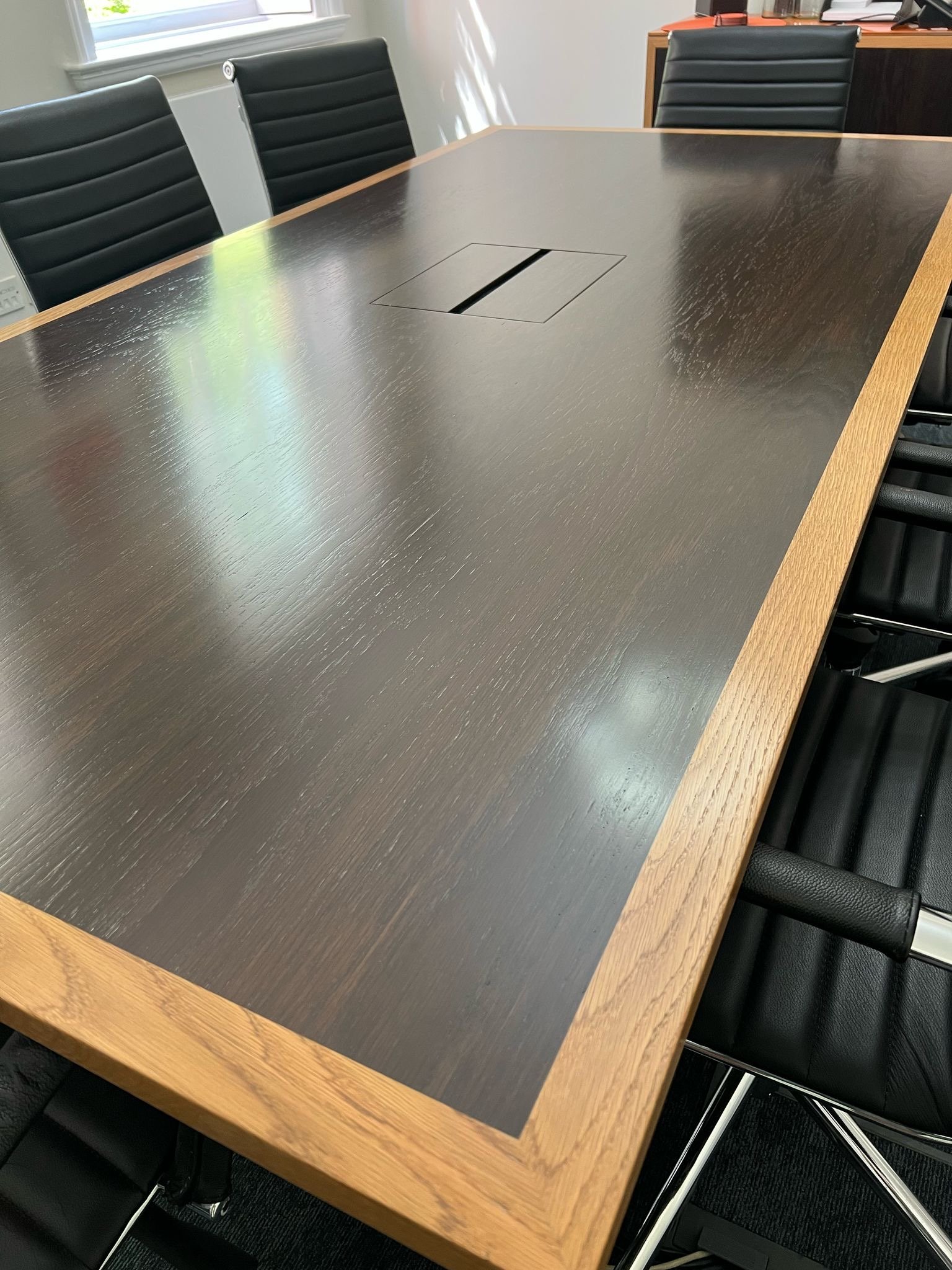 A conference table with a dark wood surface and a light wood border, surrounded by black office chairs. The table has a built-in power outlet or communication port in the center.