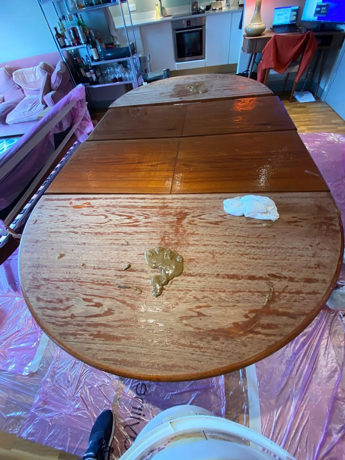 A partially stained wooden dining table with some liquid spills and tissue paper on it, set in a room with a living area and kitchen in the background.