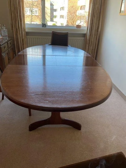 Wooden oval conference table with one black office chair, positioned in front of a window showing trees and apartment buildings outside.