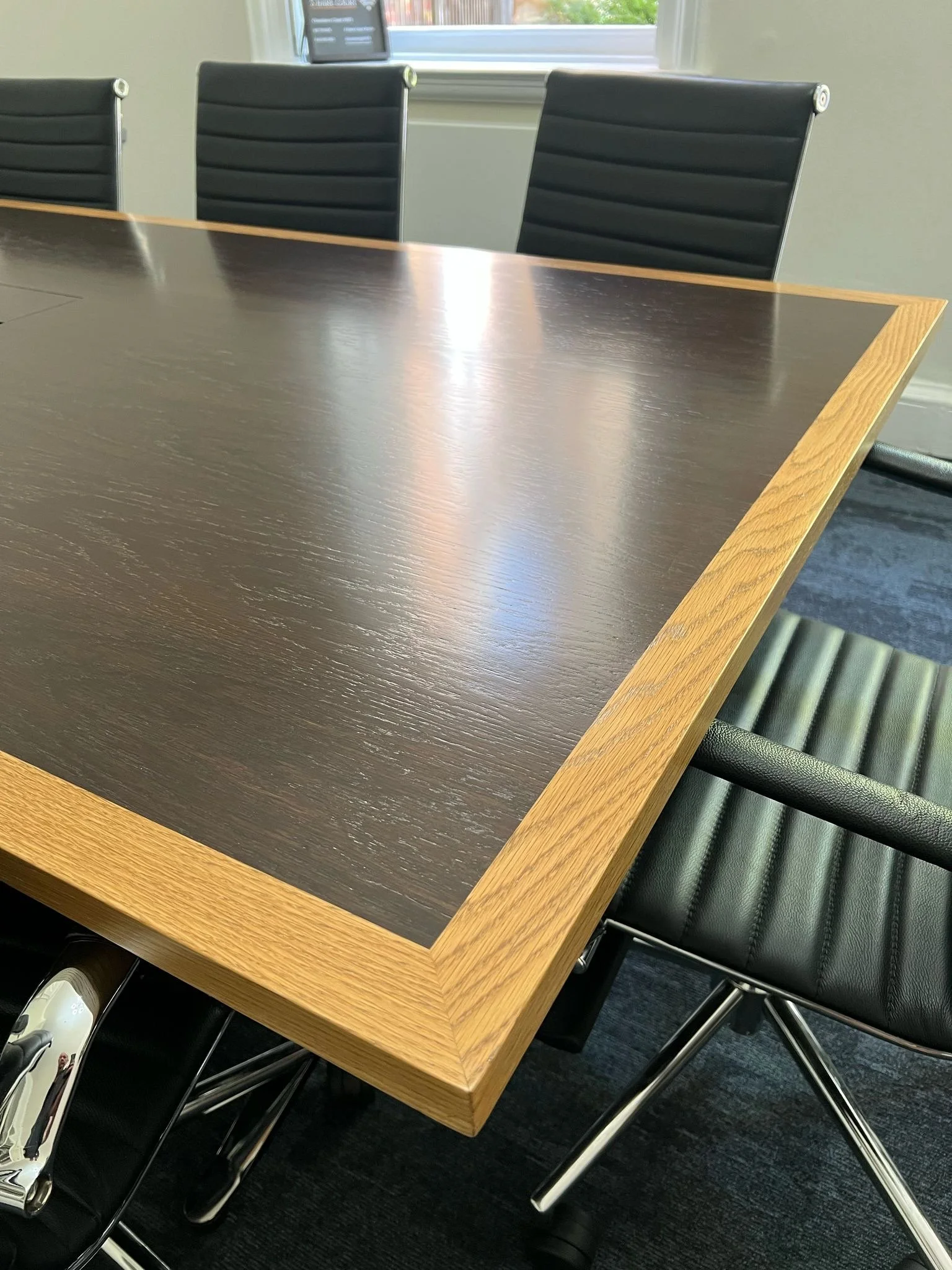 A conference room table with a dark wood surface and light wood edges, surrounded by black leather chairs with chrome legs, in front of a window.