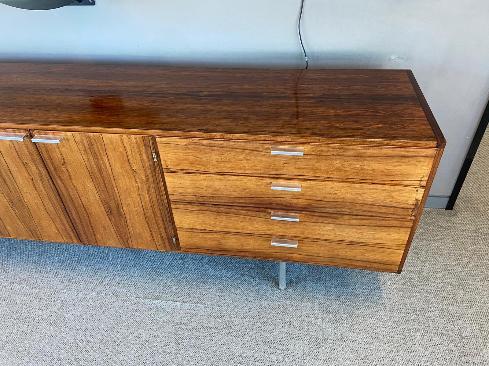 A mid-century modern wooden sideboard with four drawers and a door, metal handles, and a single metal leg, positioned on a light-colored carpet next to a gray wall with a black vertical strip on the right side.