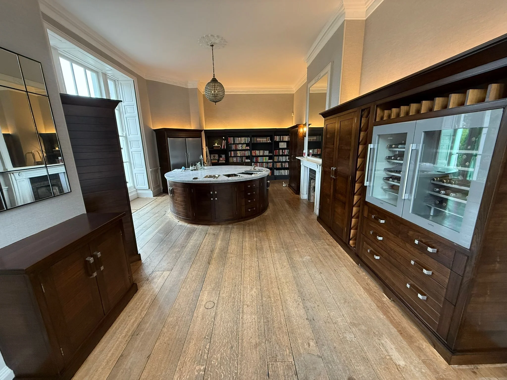 Interior view of a room with wooden flooring, dark wood furniture, bookshelves, a circular bar or island, a fireplace, a glass wine cabinet, and a window with multiple panes.