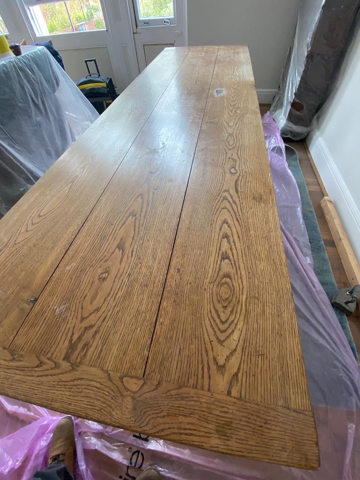 A wooden dining table with a natural finish sitting on a plastic-covered floor in a sunlit room.