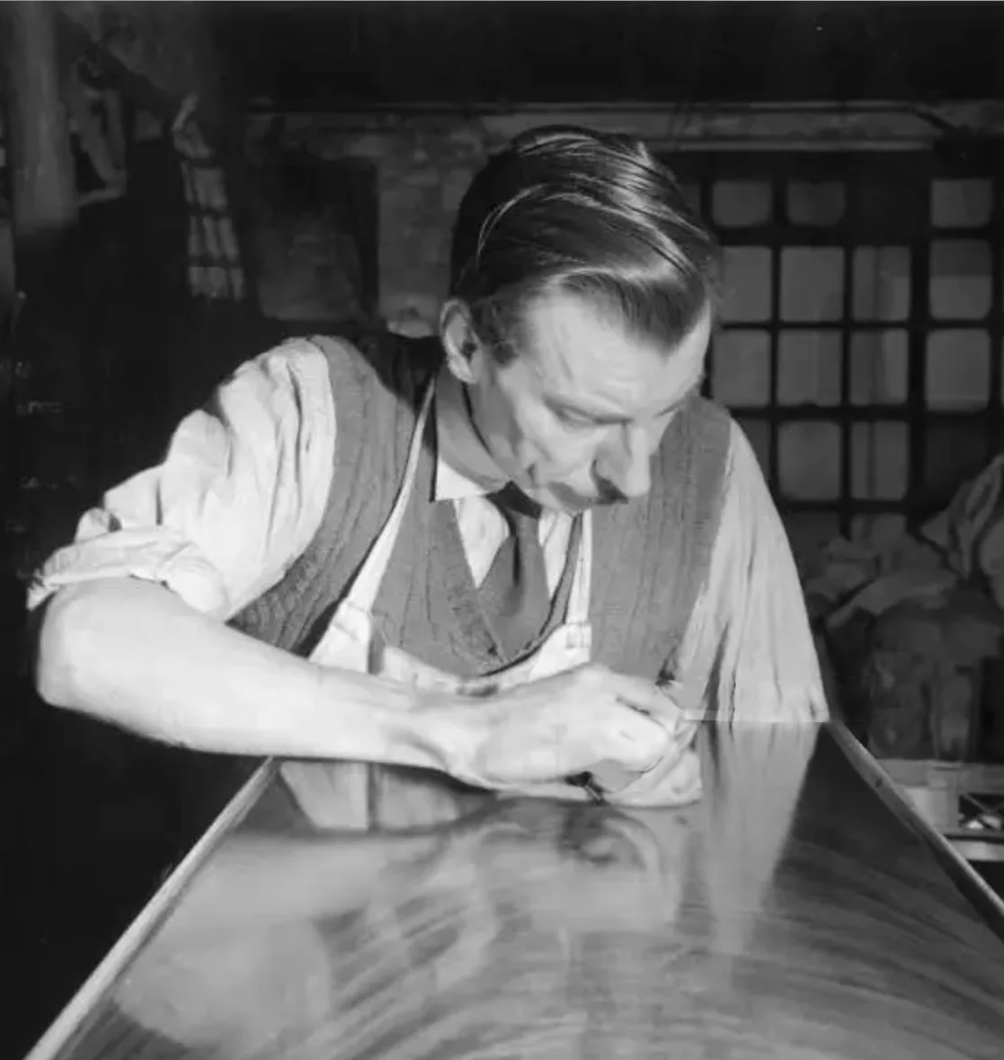 Black and white photo of a man with slicked-back hair and a mustache, wearing a vest, shirt, and tie, working on a polished wooden surface, possibly a table or piece of furniture, in a workshop setting.