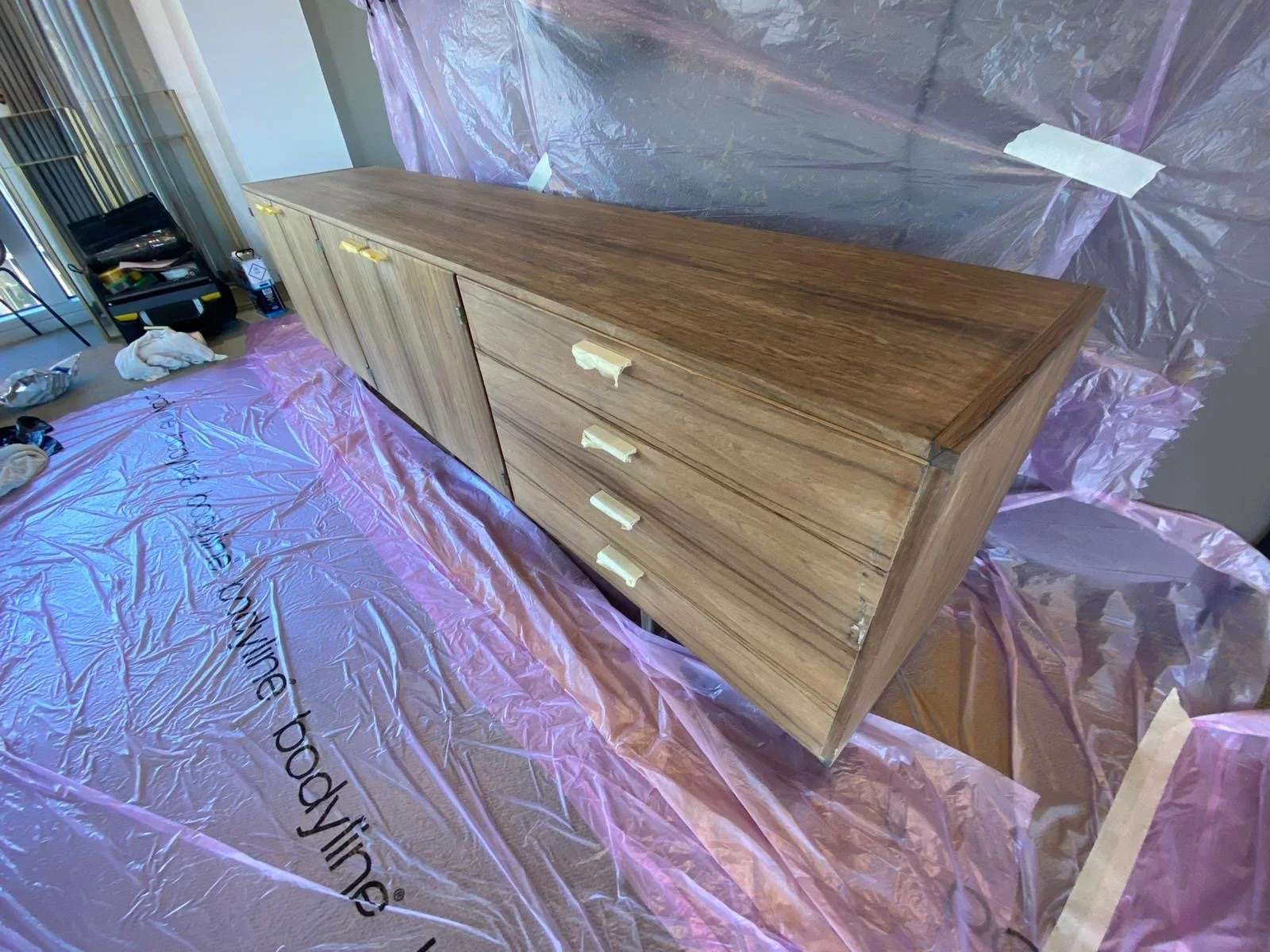Wooden sideboard with four drawers and a cabinet, placed on a surface covered with pink plastic sheeting in a room under renovation.