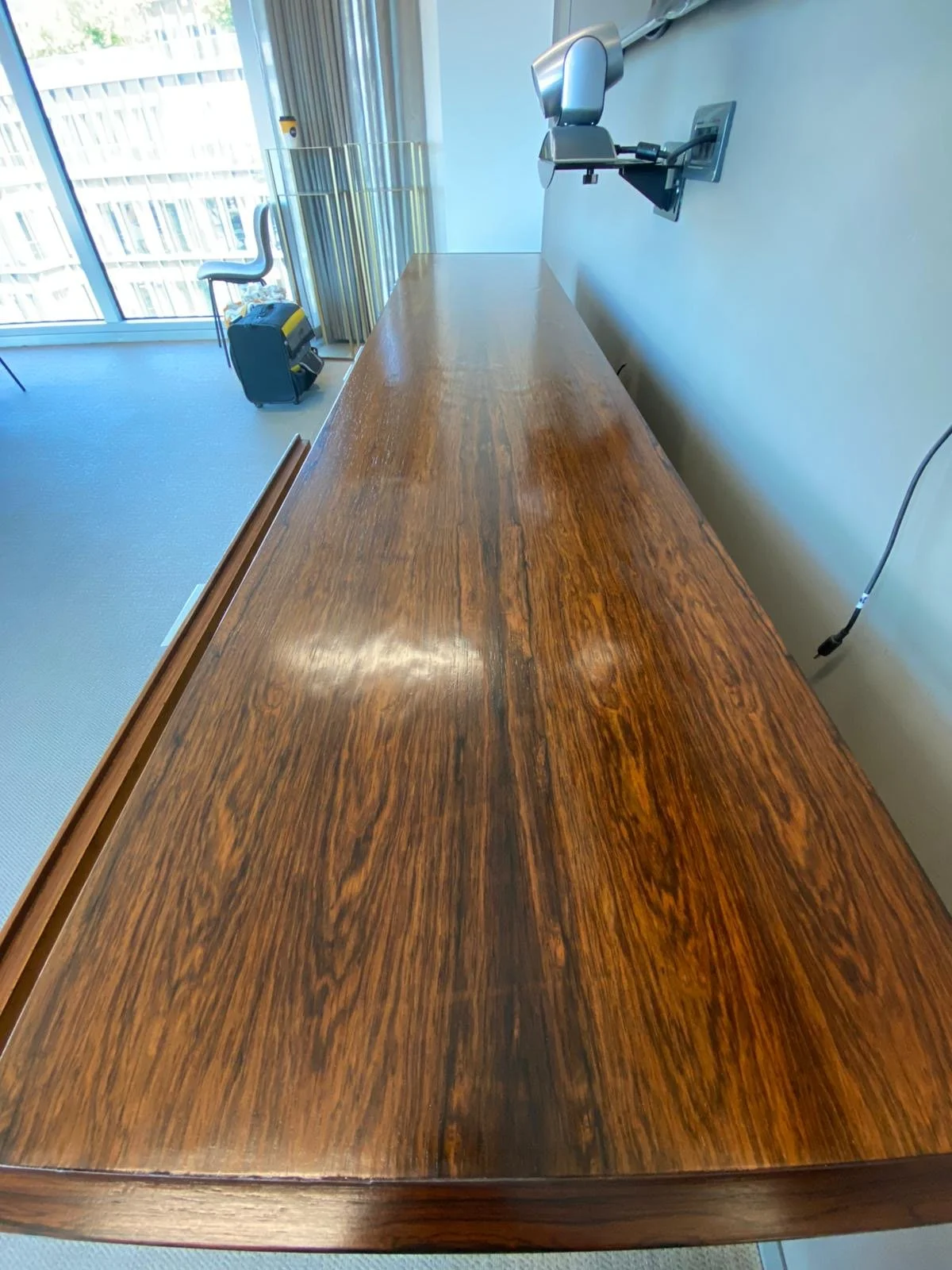 A long wooden conference table in a modern office with large windows and a view of a building's exterior, with some chairs and equipment visible.