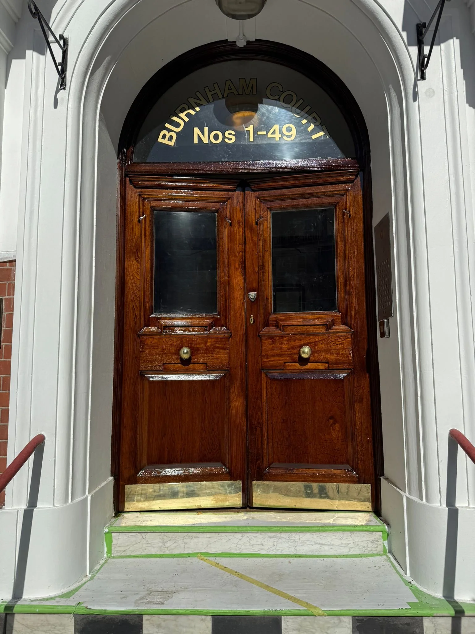Dark brown wooden double door with glass panels, located under an arched window with the address 'Nos 1-49' and 'BURNHAM COUNTY' written on it, with white trim and steps leading up to the entrance.