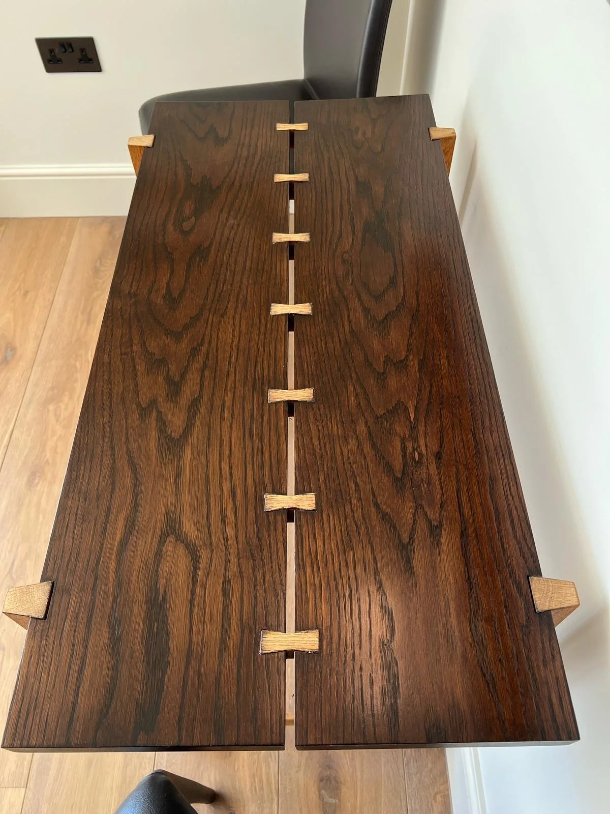 Close-up of a wooden table with a central split and small wooden wedge inserts along the split, in a room with a power outlet on the wall.