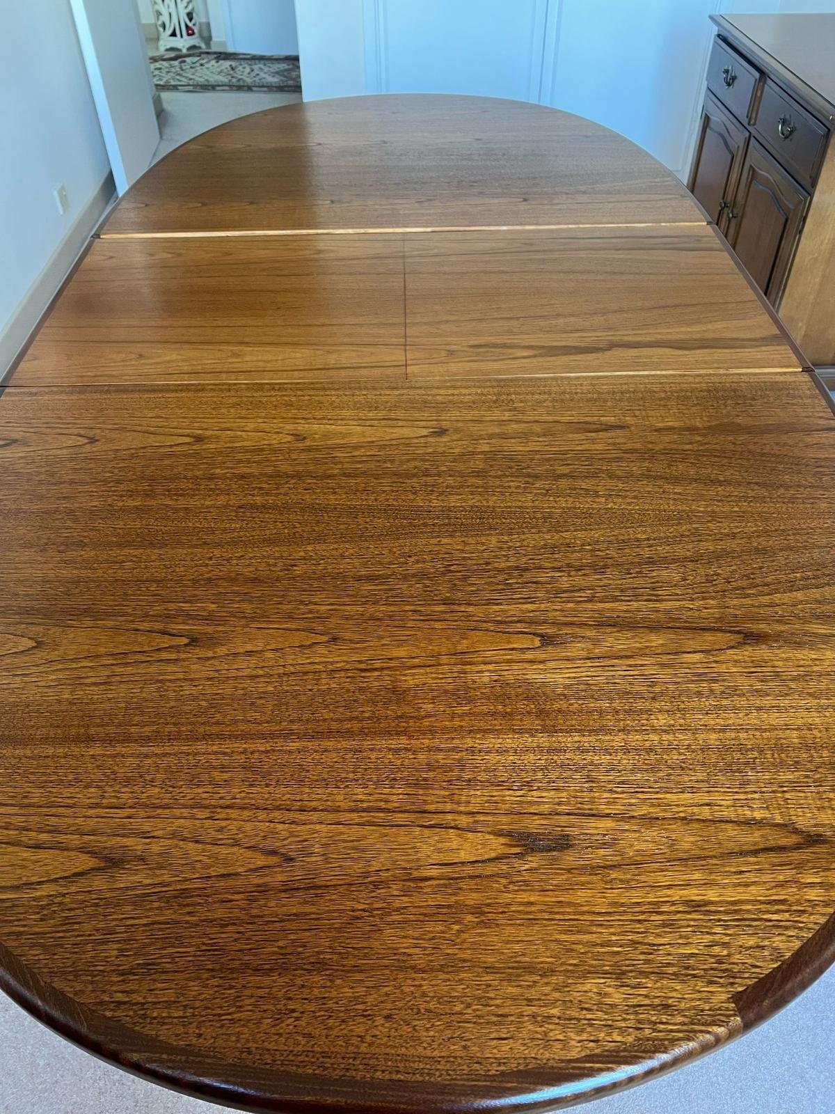A wooden dining table with a polished surface and a rounded edge, situated in a room with a cabinet and a door in the background.
