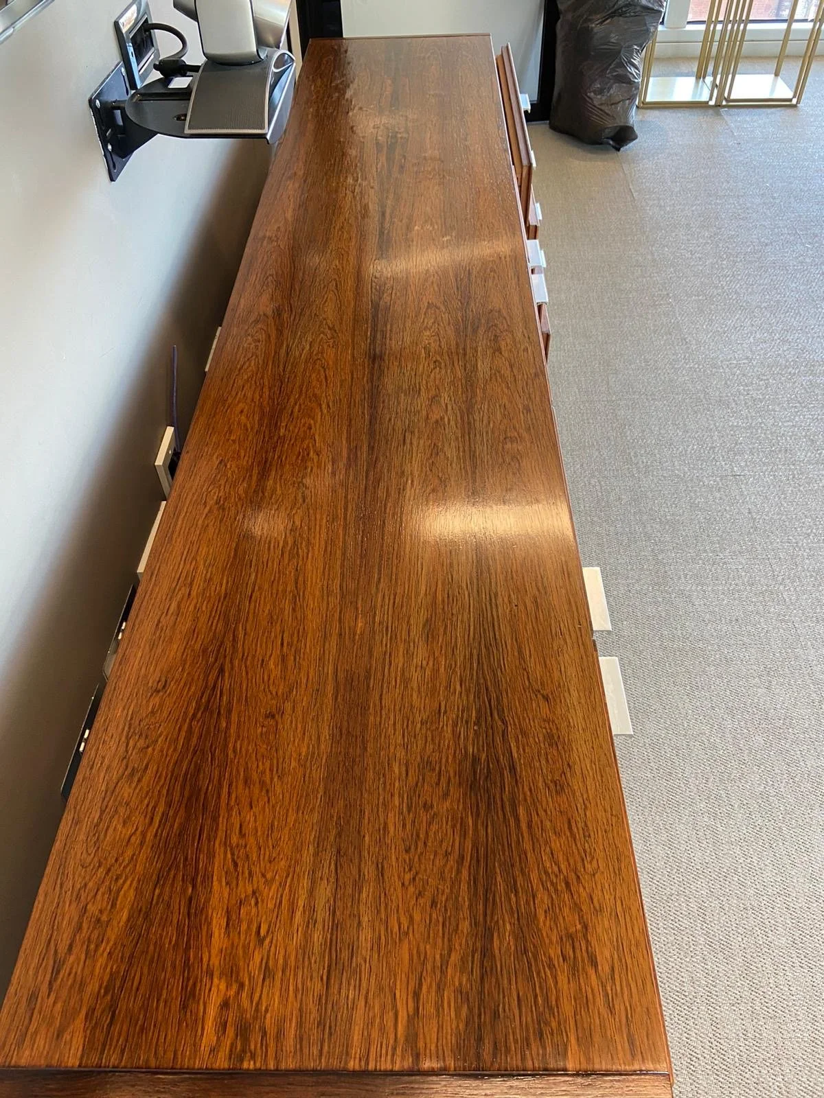 A long wooden conference table in a room with gray carpet, a trash bag, and gold metal shelving to the side, with a room divider visible in the background.
