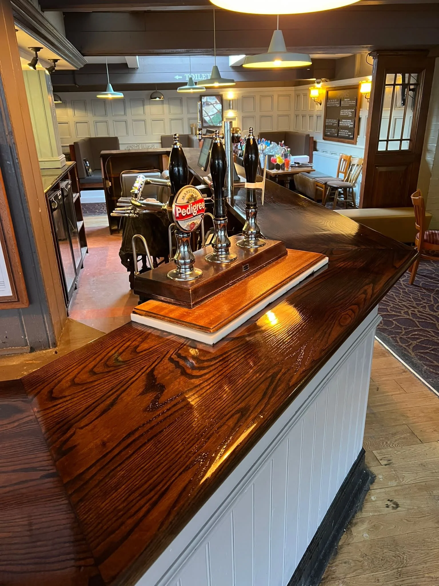 View of a bar counter with beer taps, inside a restaurant with dim lighting, booths, chairs, and a table with drinks and bottles in the background.
