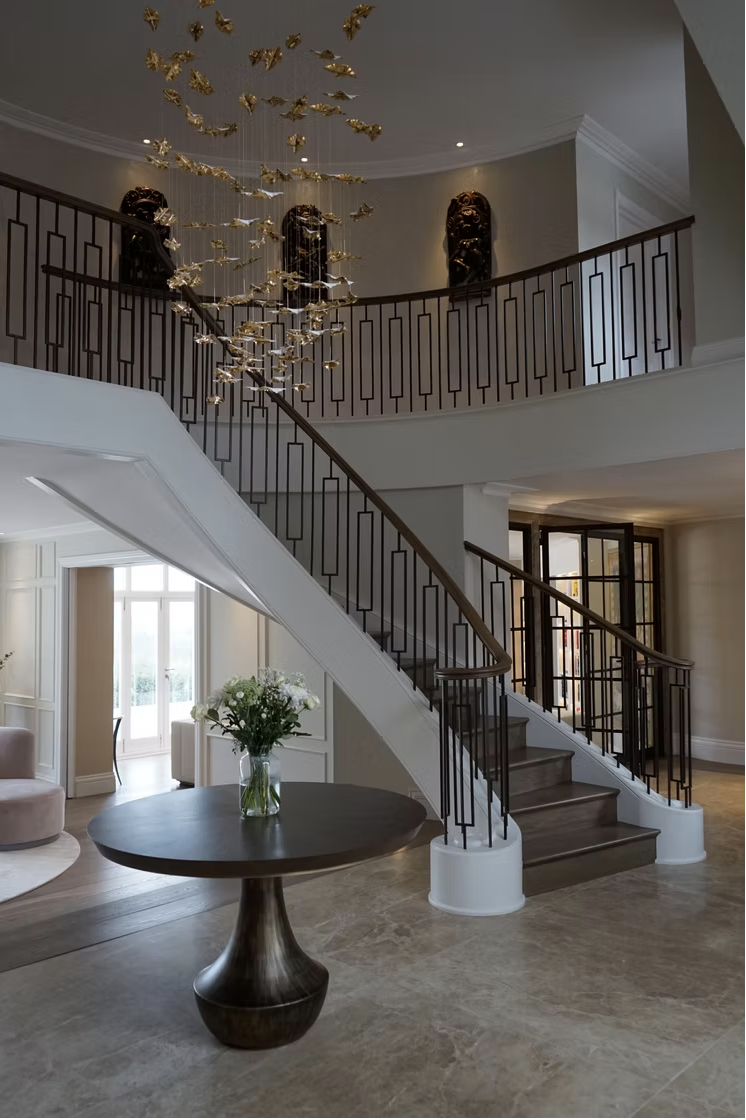 Interior view of a grand staircase in a luxurious home with a modern chandelier hanging from the ceiling, a round dark wood table with a flower arrangement, and a balcony with decorative sculptures.