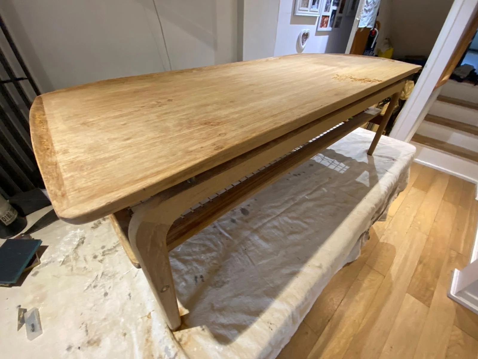 Unfinished wooden table with a natural finish placed on a covered surface in a room with wooden flooring and stairs in the background.