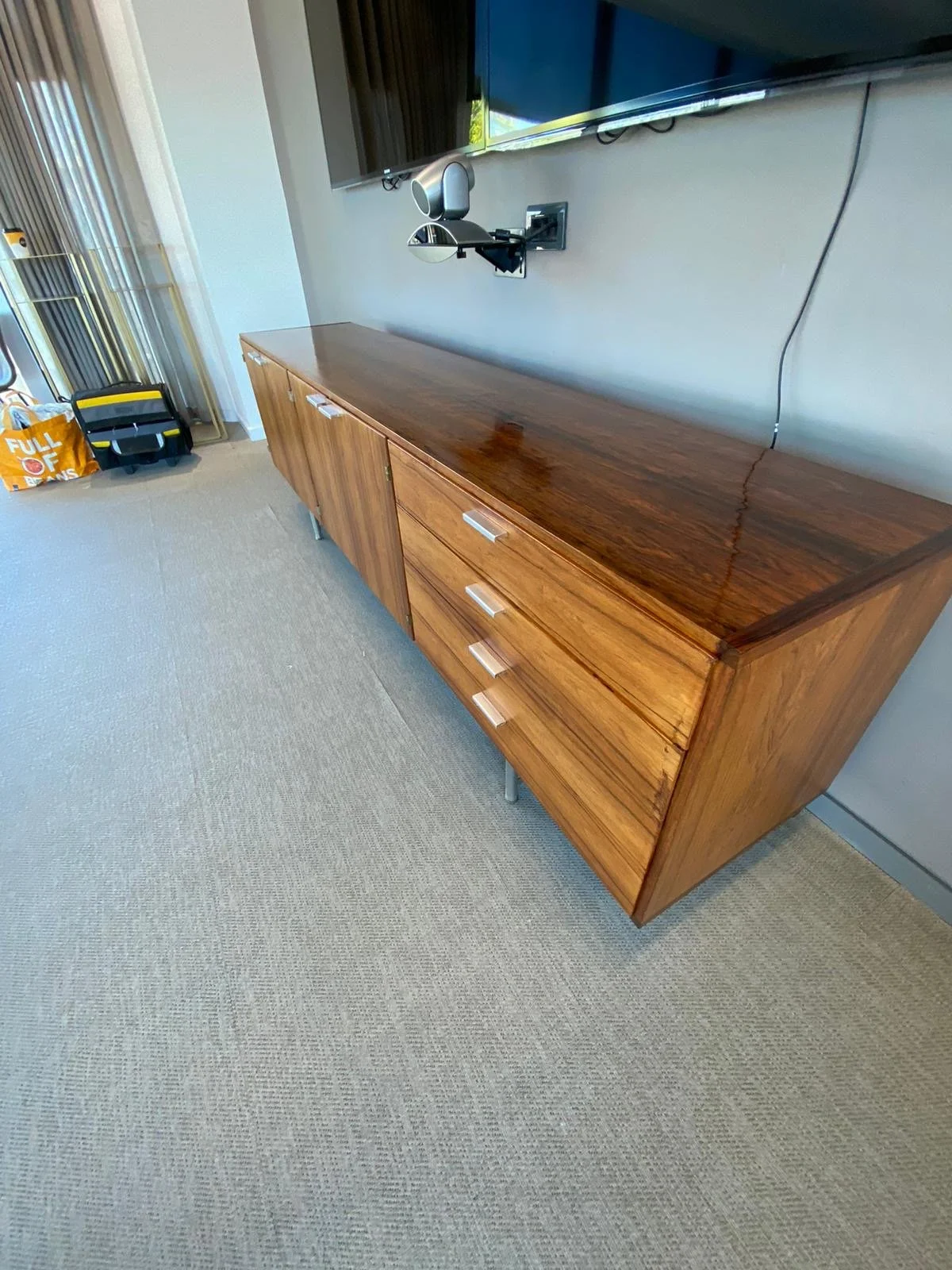 A wooden sideboard with three drawers and two cabinets, mounted on metal legs, placed against a wall with a television mounted above it.