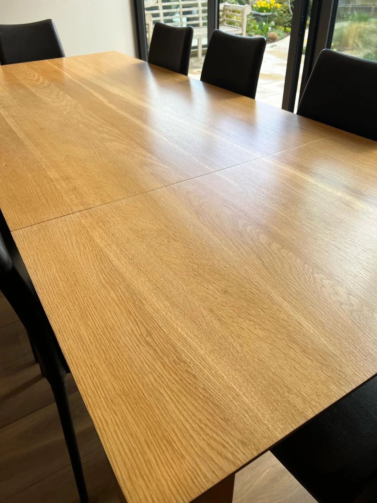 A wooden dining table with black chairs around it, positioned near large glass doors overlooking a patio with greenery and yellow flowers.