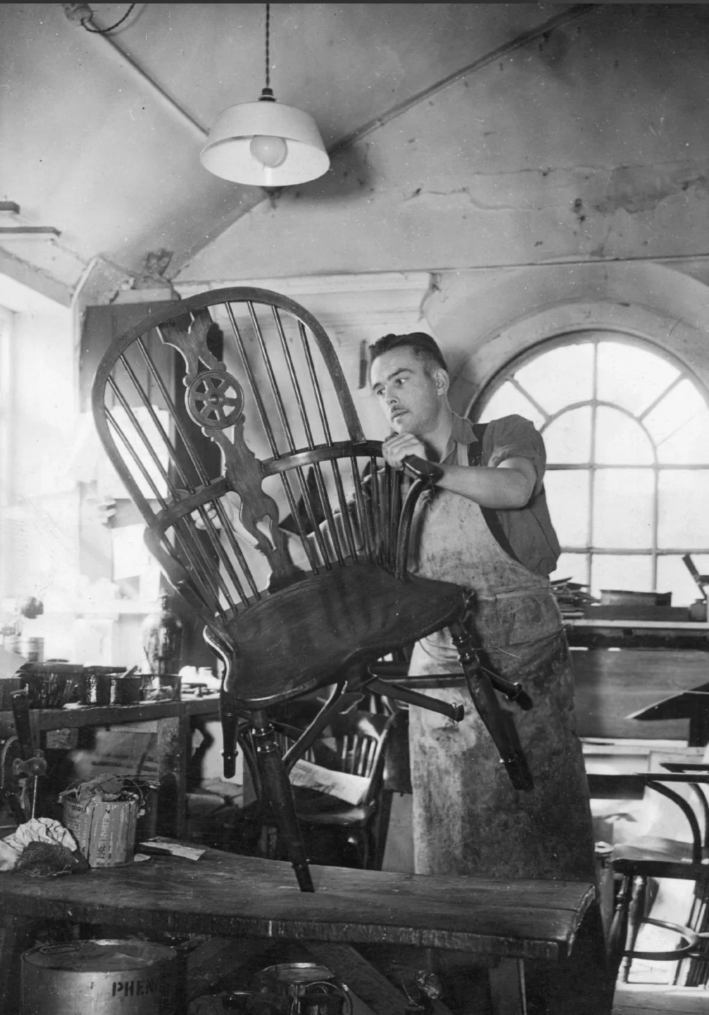 A man working to restore a wooden chair in a workshop, with tools and furniture around, and a large window in the background.