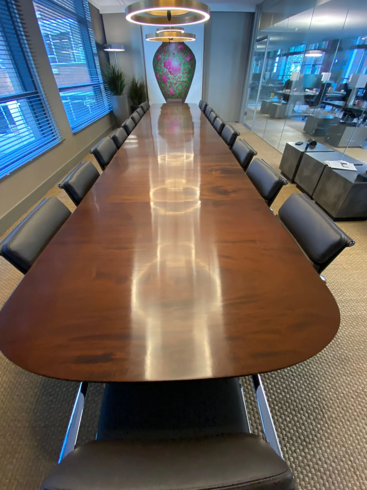 A long wooden conference table with sixteen black chairs, eight on each side, in a modern office meeting room. The room has large windows with blinds, a vase and plants at the far end, and a glass wall to an adjoining office area.
