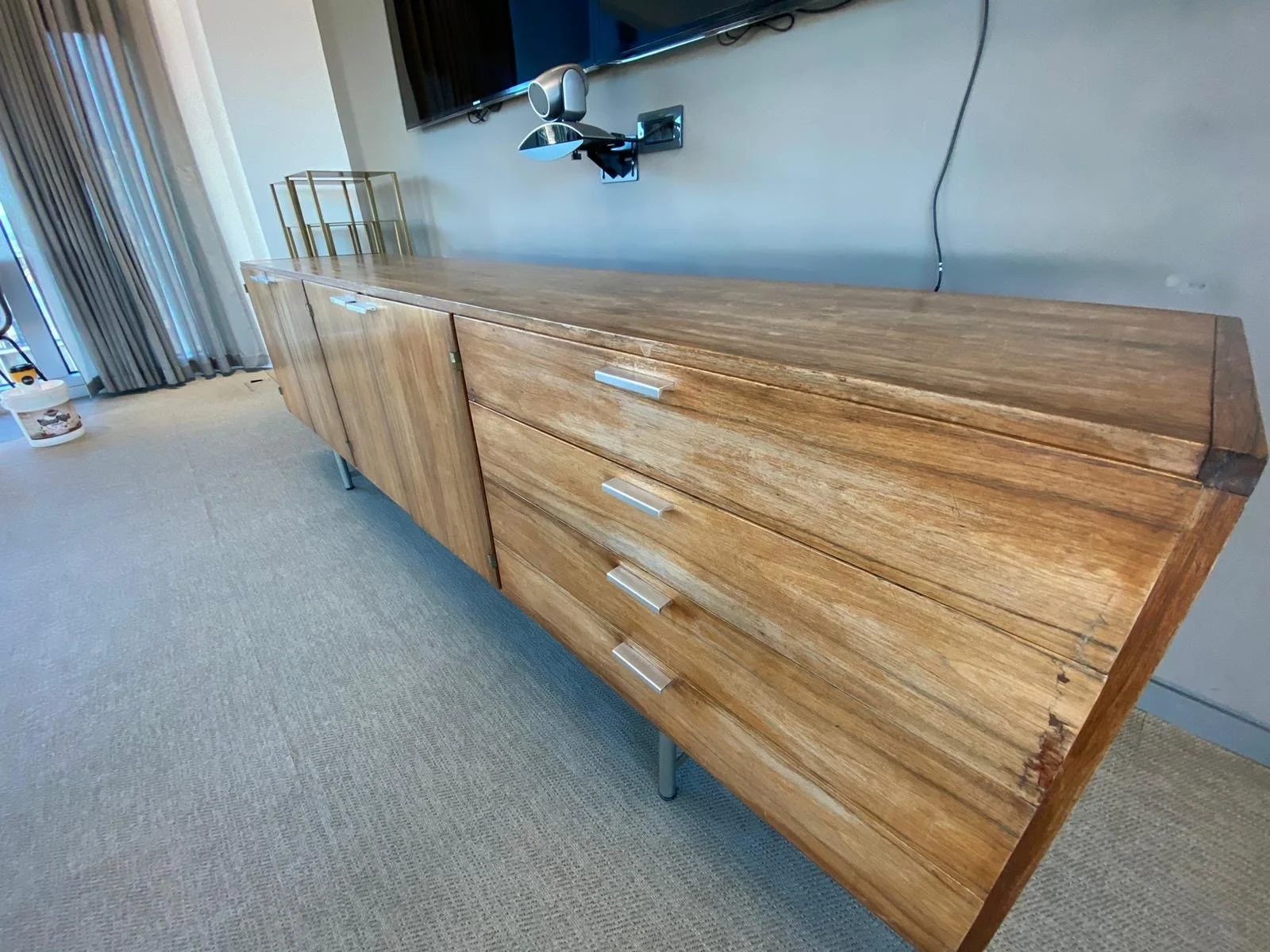 A long wooden sideboard with drawers and cabinets, positioned against a gray wall with an electronic device mounted above. The room has beige carpet flooring and curtains near a glass sliding door.