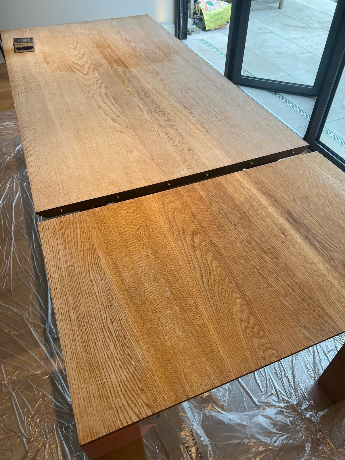 Two wooden tabletops placed side by side near a window, with a small tape measure on the corner of one table, and a plastic sheet on the floor underneath.