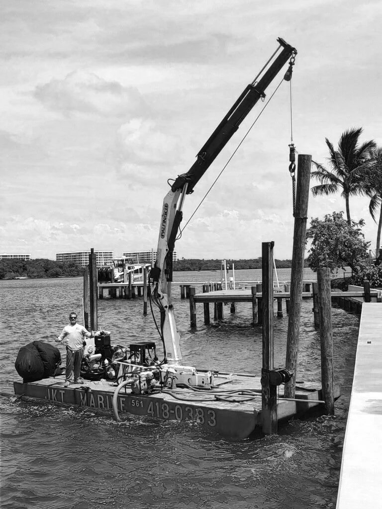 Small work boat with crane near dock, man standing on deck, palm trees in background