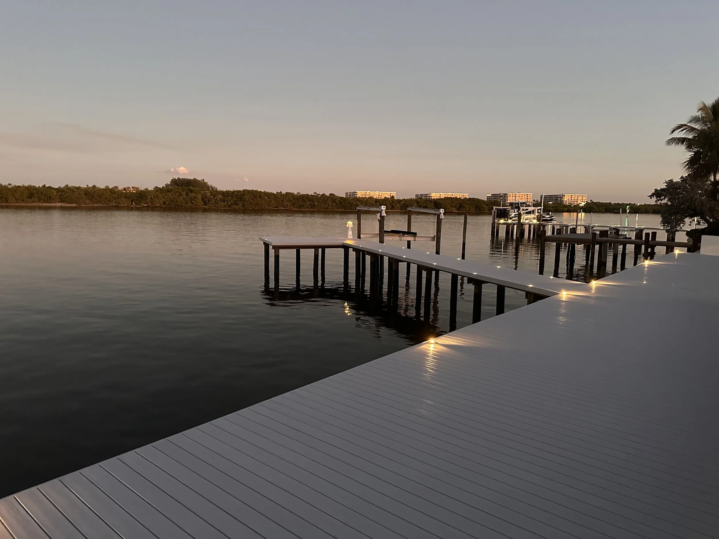 A serene waterfront scene at dusk with a wooden pier extending into a calm body of water. The pier and nearby piers have illuminated lights. A palm tree is visible on the right and buildings are in the background across the water.