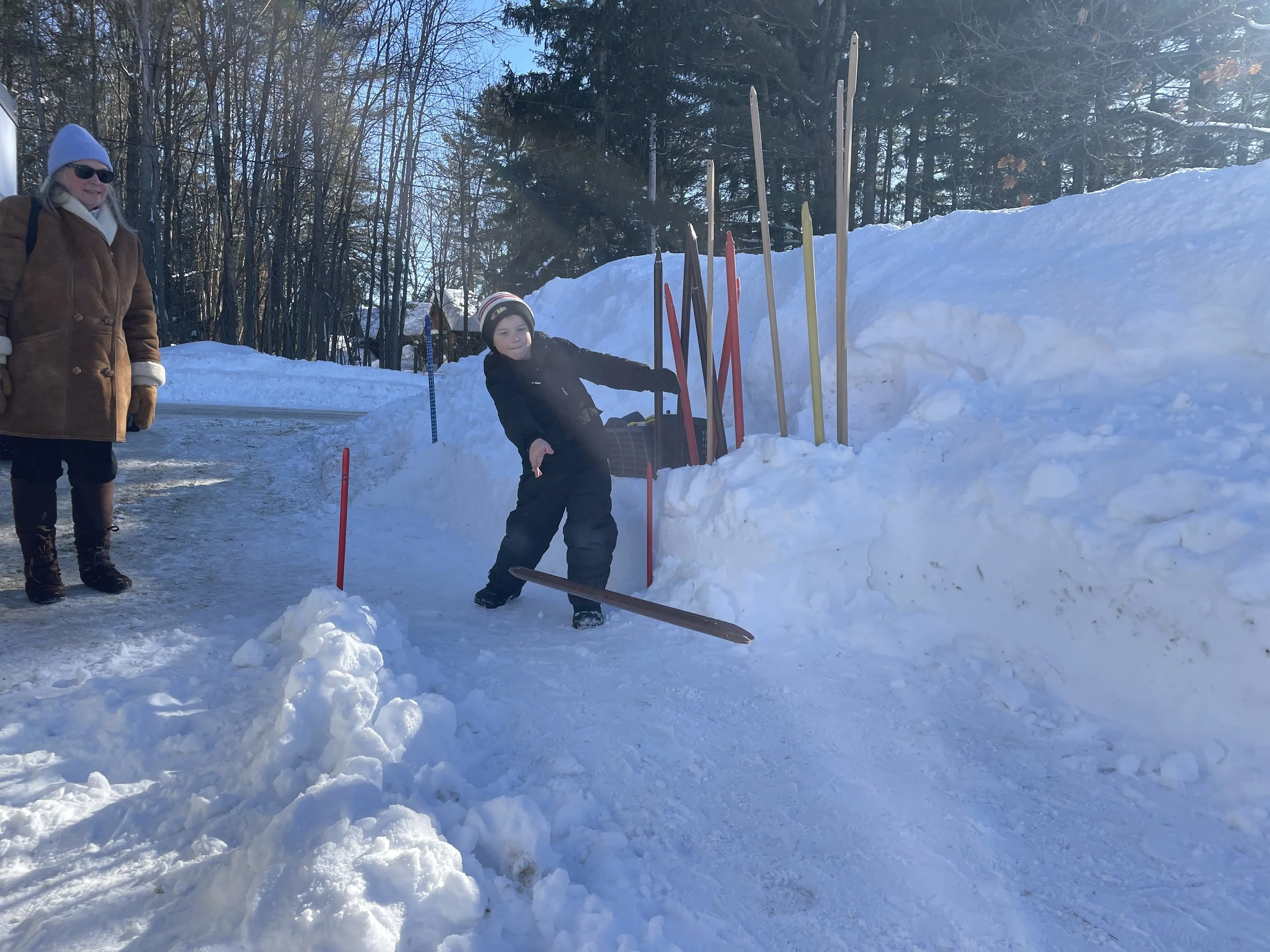 A young boy in winter clothing sliding on a snow-covered ice rink with an adult woman watching nearby. Snow piles and wooden sticks are seen in the background.