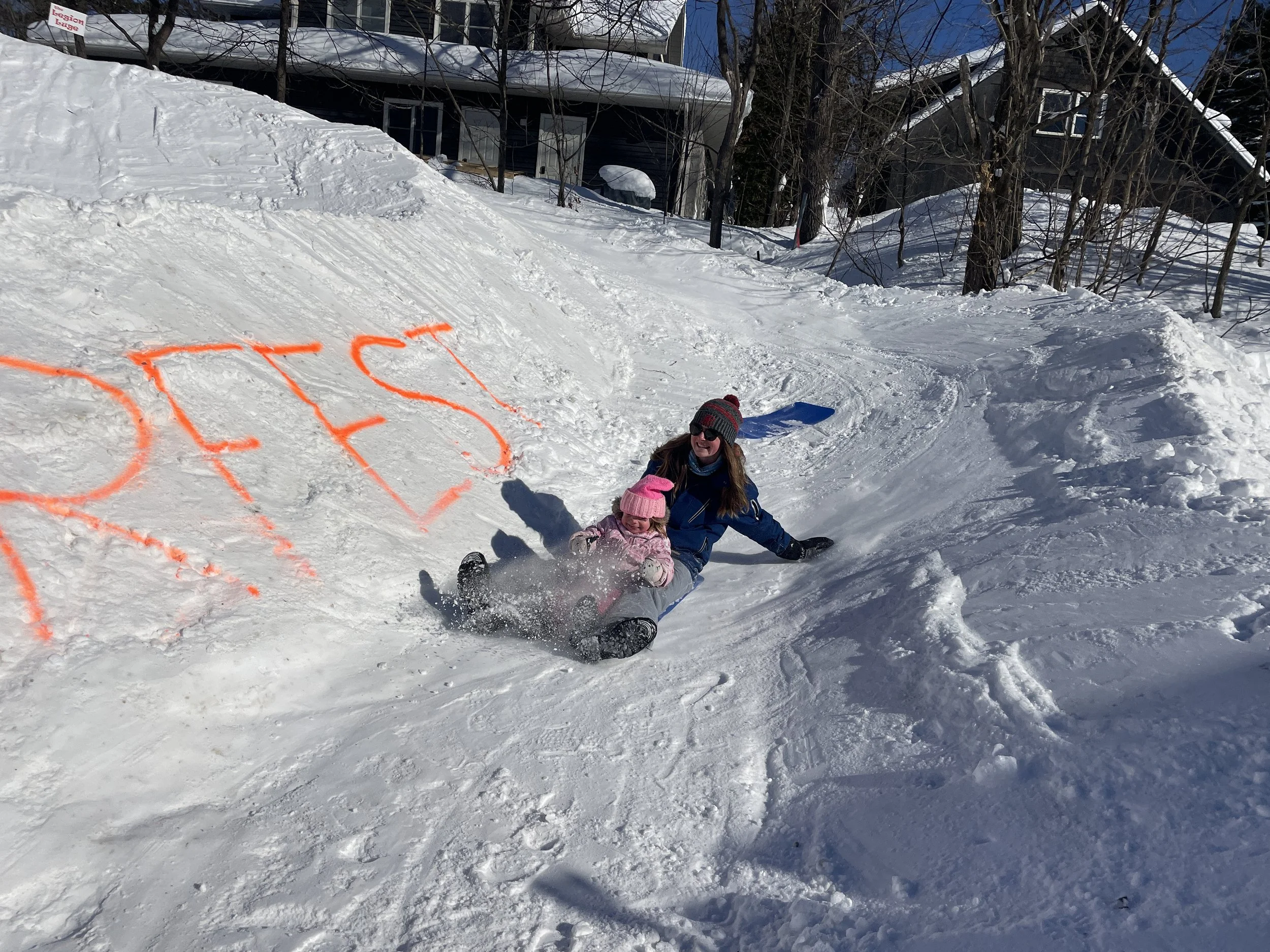 A woman and a young girl in winter clothing sliding down a snowy slope on a small snowboard or sled, with trees and houses in the background and the word 'PEACE' spray-painted in orange on the snow.