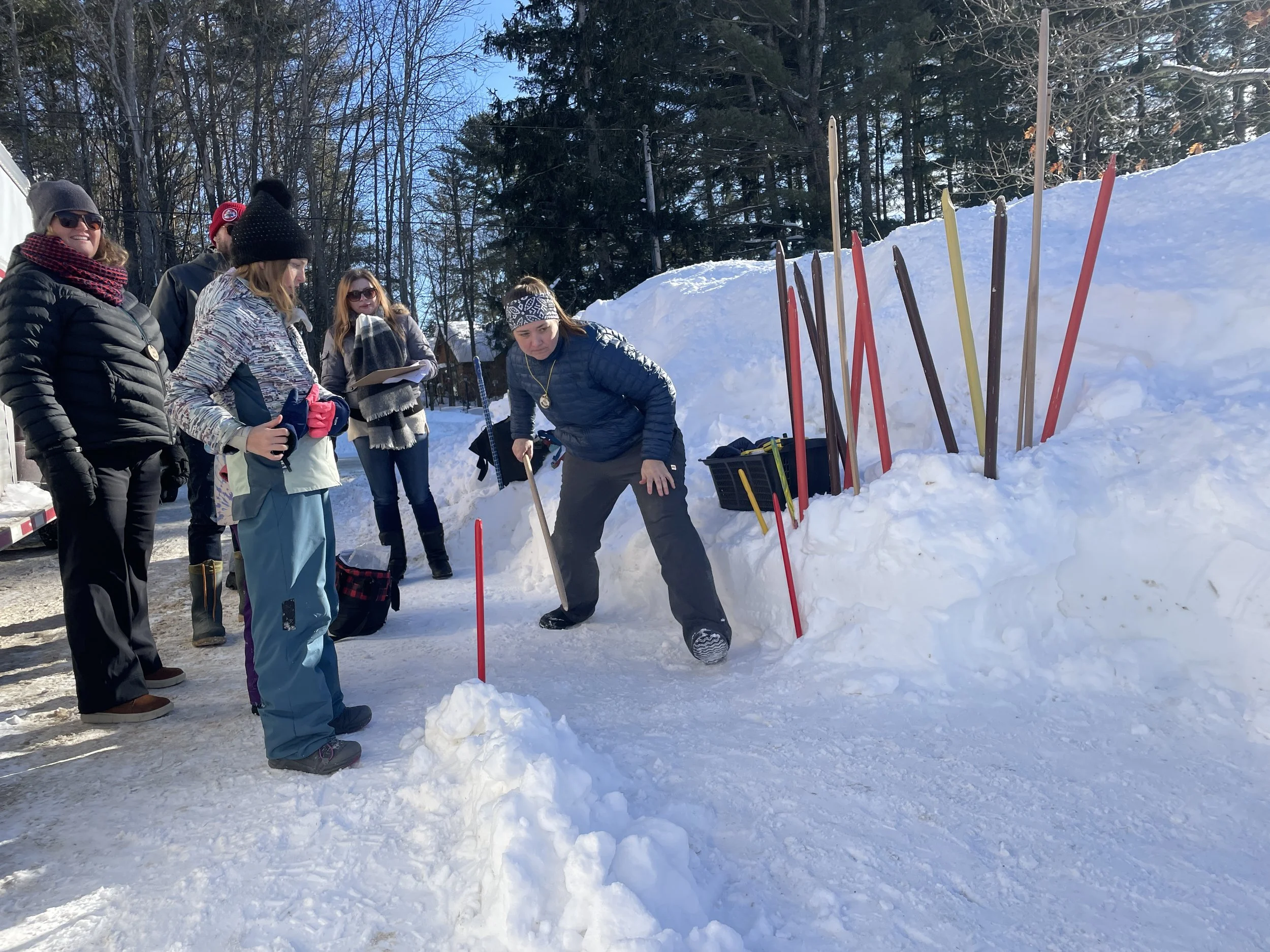 A group of six people dressed warmly in winter clothing, gathered outdoors in snowy conditions, observing a woman demonstrating snow shoveling techniques next to a pile of snow with colored sticks protruding from it.
