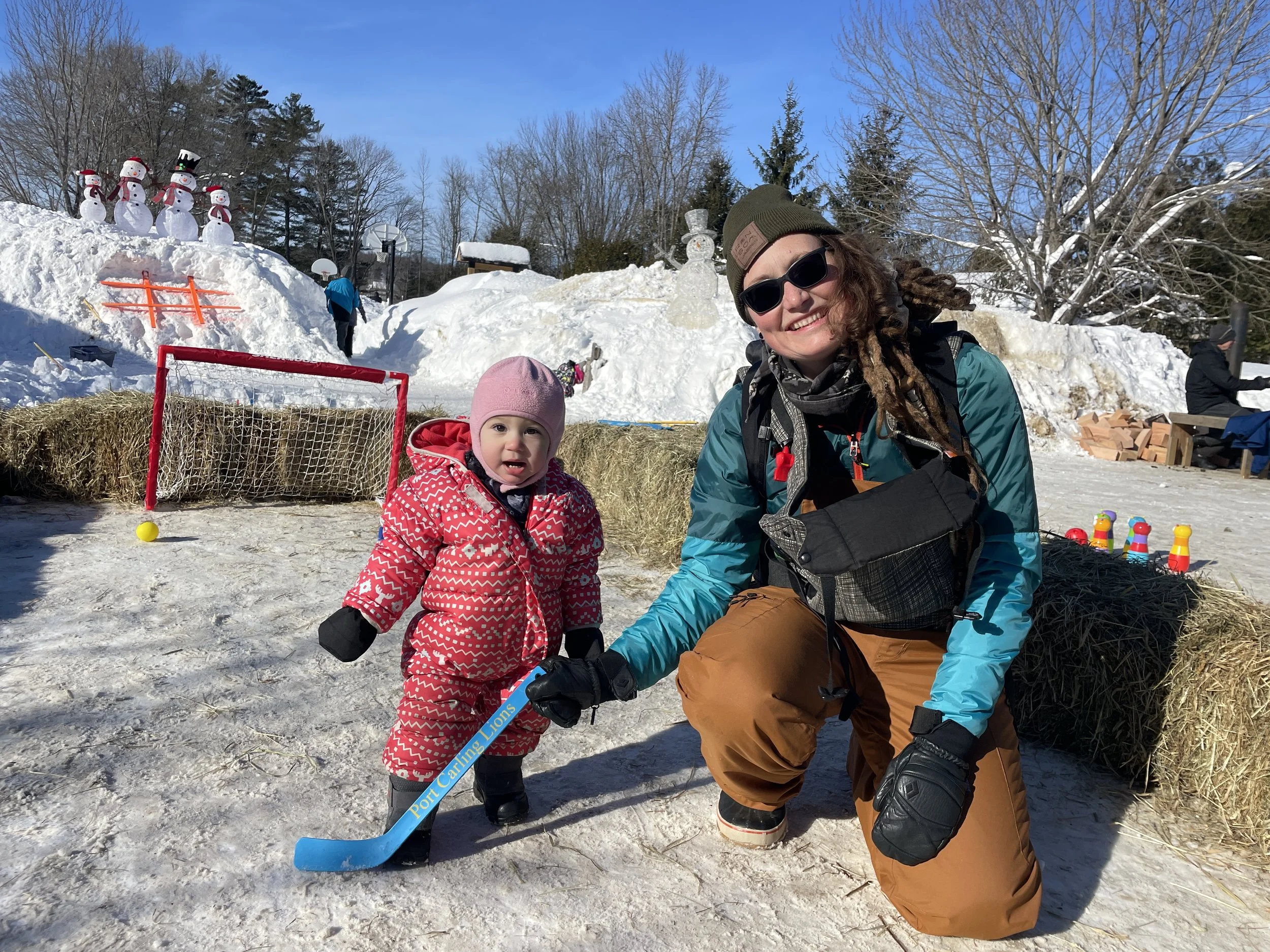 A woman with sunglasses, a beanie, and winter gear kneeling next to a young girl in a pink snowsuit holding a small hockey stick outdoors in a snowy area with snowmen and hay bales in the background.