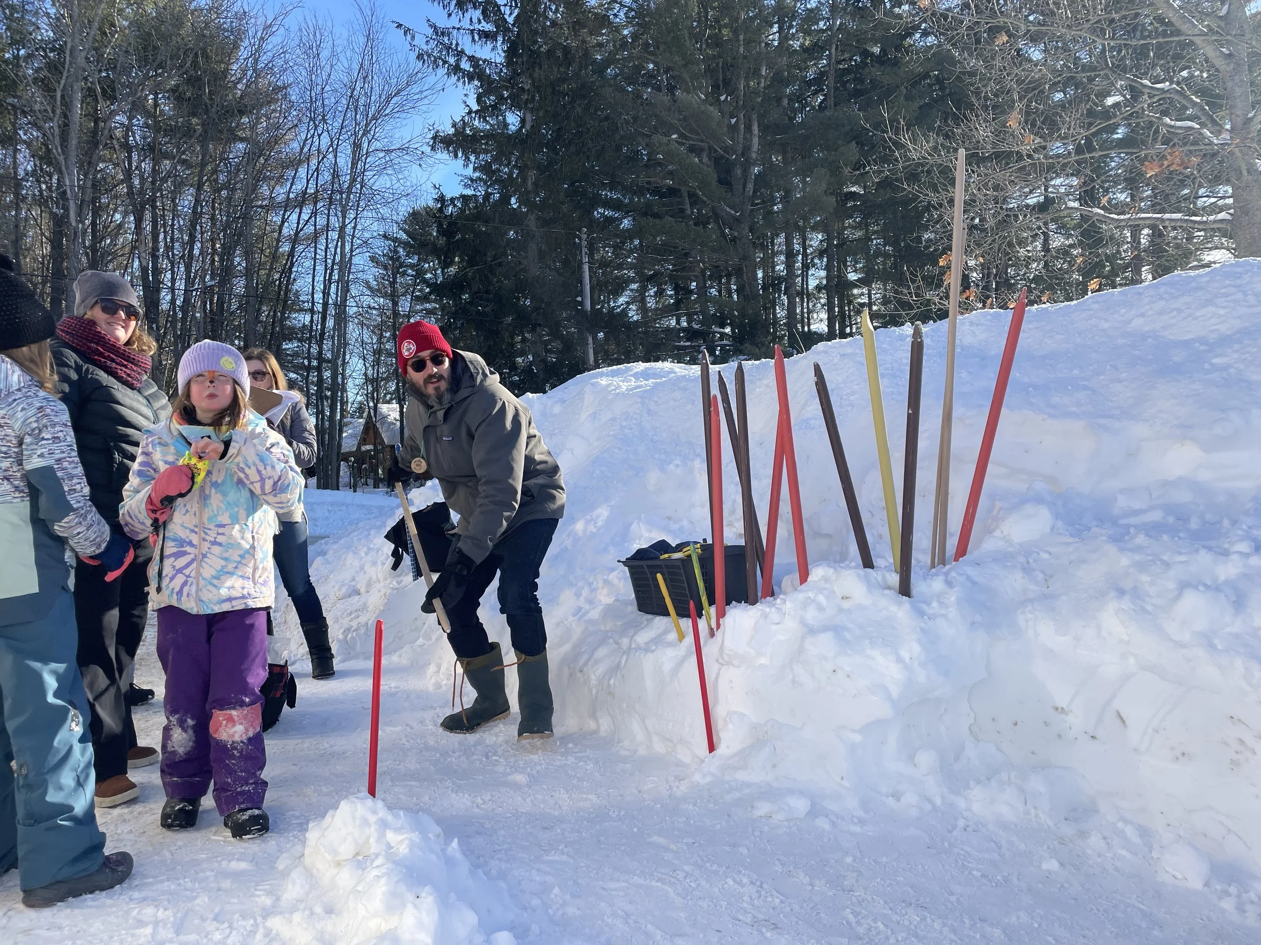 Group of people outdoors in the snow, gathered around with snow shovels and colorful sticks nearby, in a winter scene with trees and a snowbank.