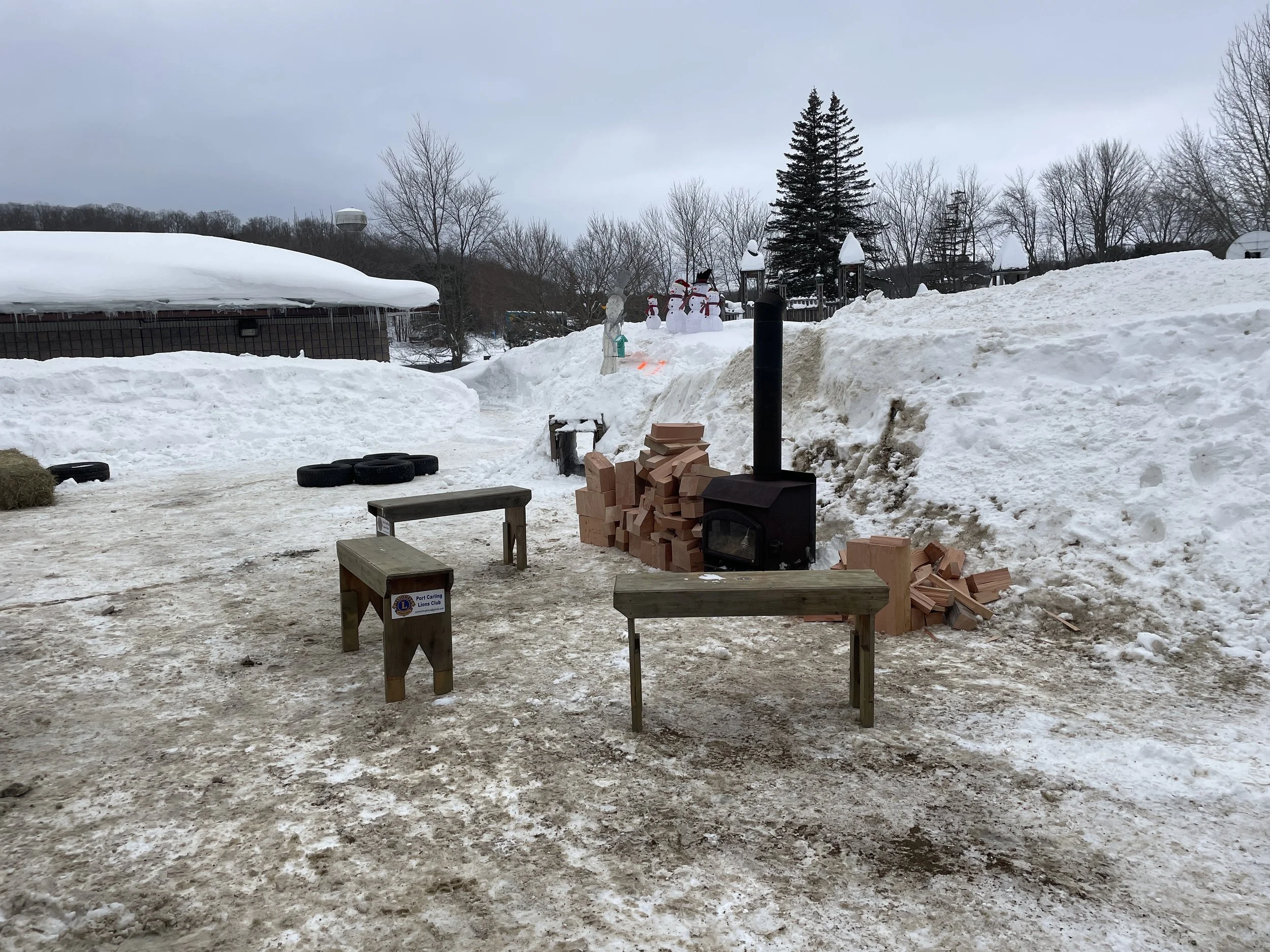 Outdoor scene with snow-covered ground, a wood-burning stove with a chimney, stacks of firewood, benches, tires, and snowmen in the background.