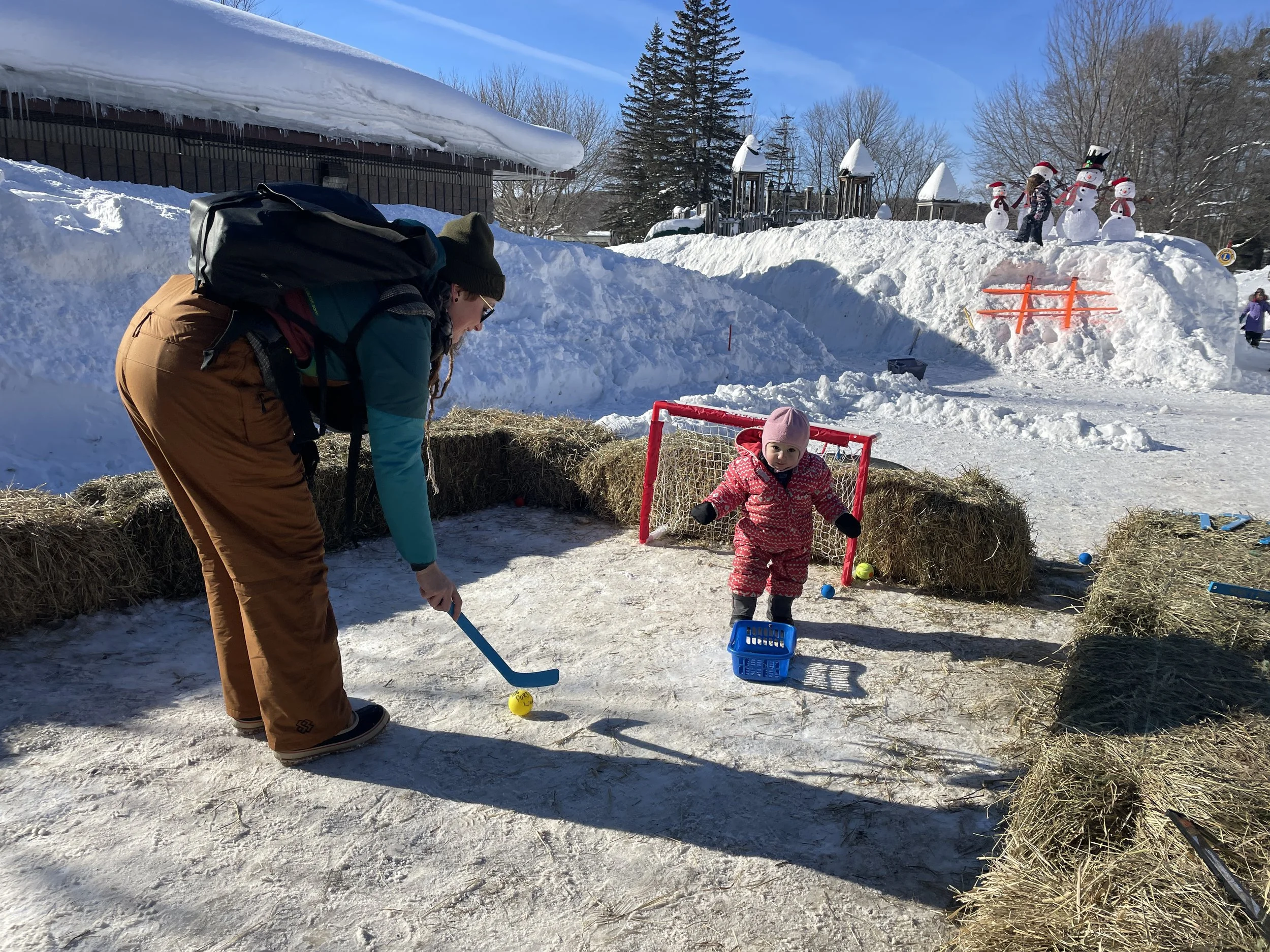 A man and young girl playing hockey outdoors in the snow. The man is bending over to hit a yellow puck, wearing winter gear, including a backpack. The girl, dressed in pink with a matching hat, stands near a small goal with a blue basket, surrounded 