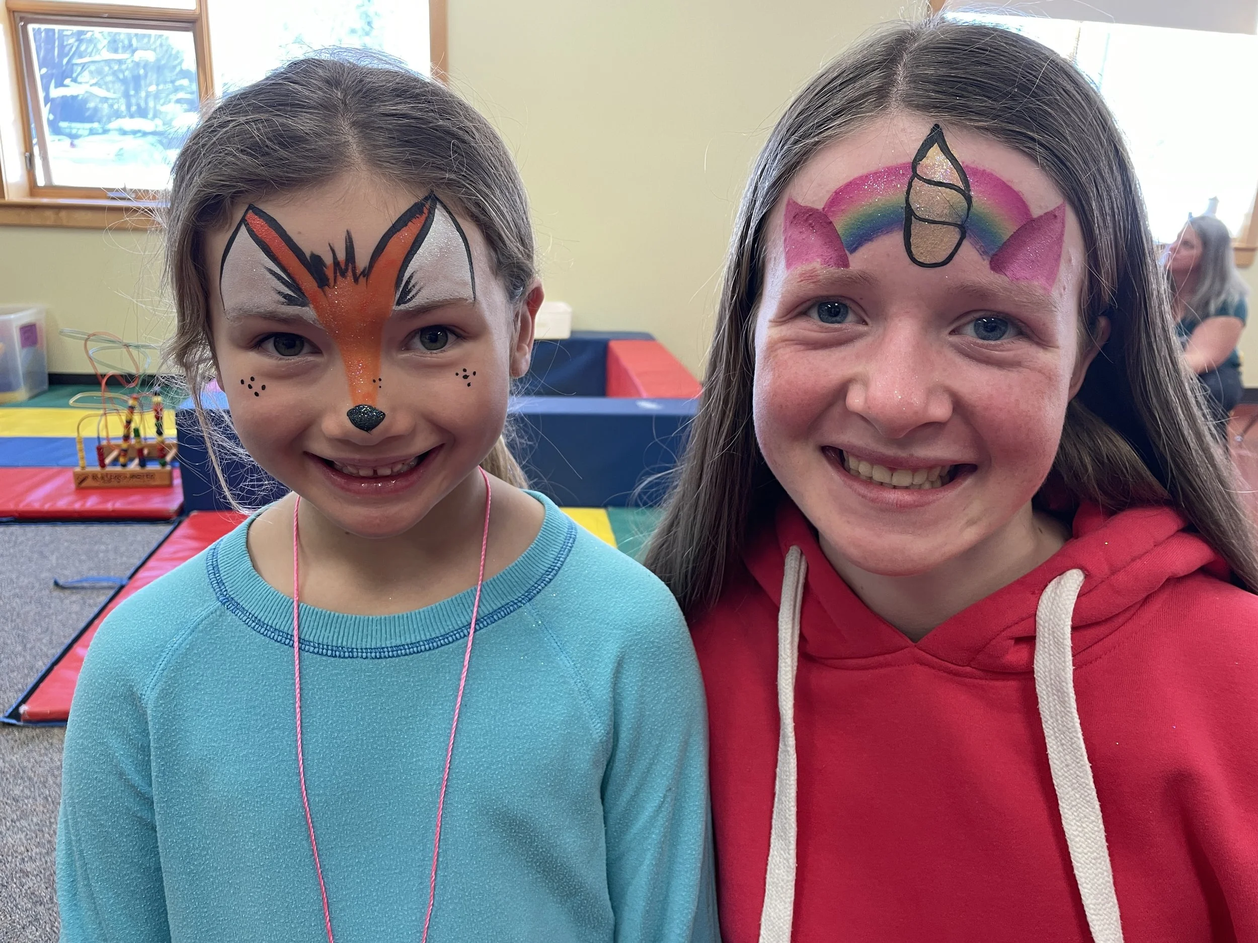 Two young girls with face paint, smiling inside a colorful indoor play area. The girl on the left has a fox face design, and the girl on the right has a unicorn face design.