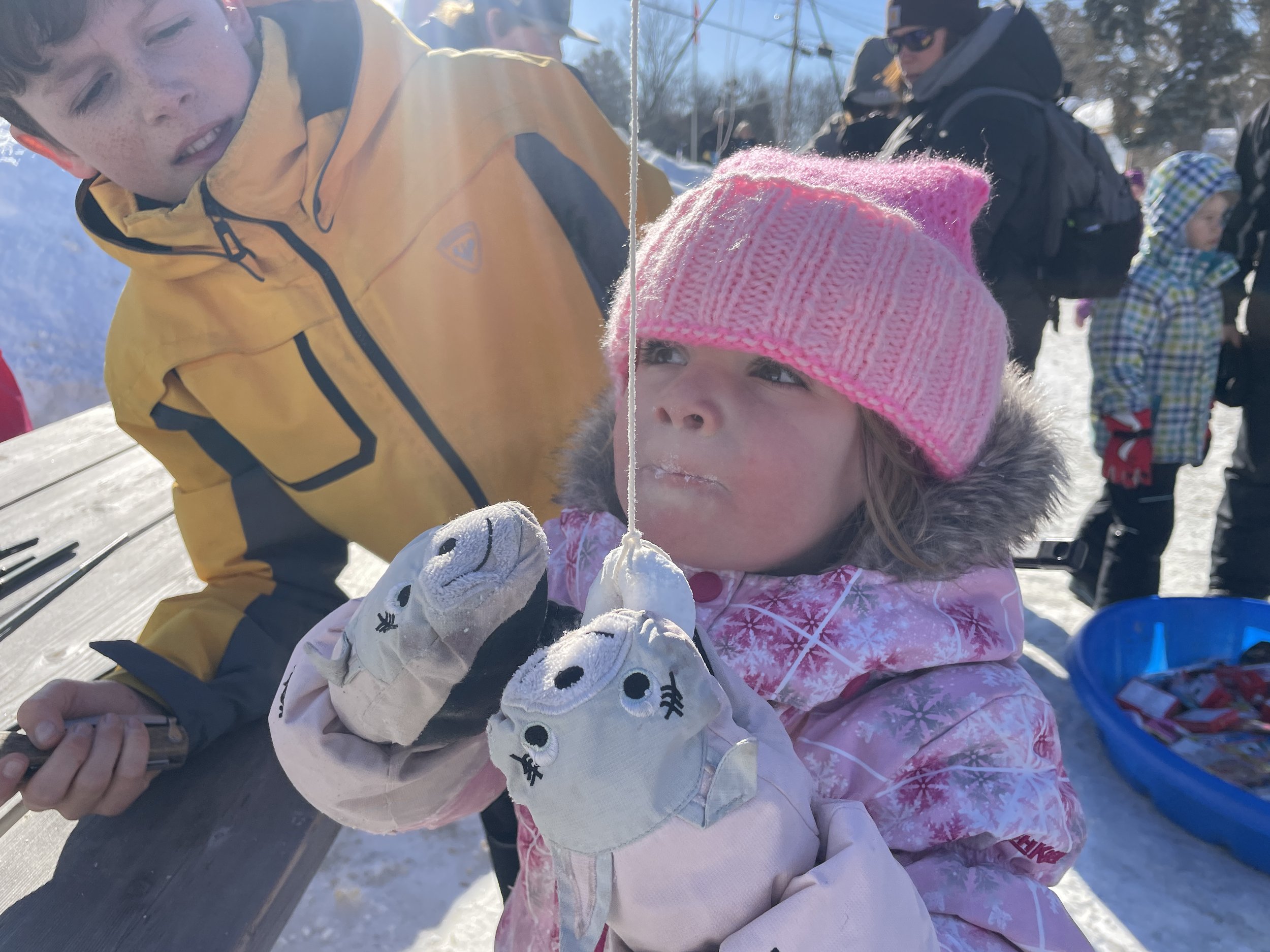 A young girl in a pink knit hat and winter coat holds a fishing line with a small fish on it, while a young boy in a yellow jacket looks on, outdoors in a snowy area with other people in the background.