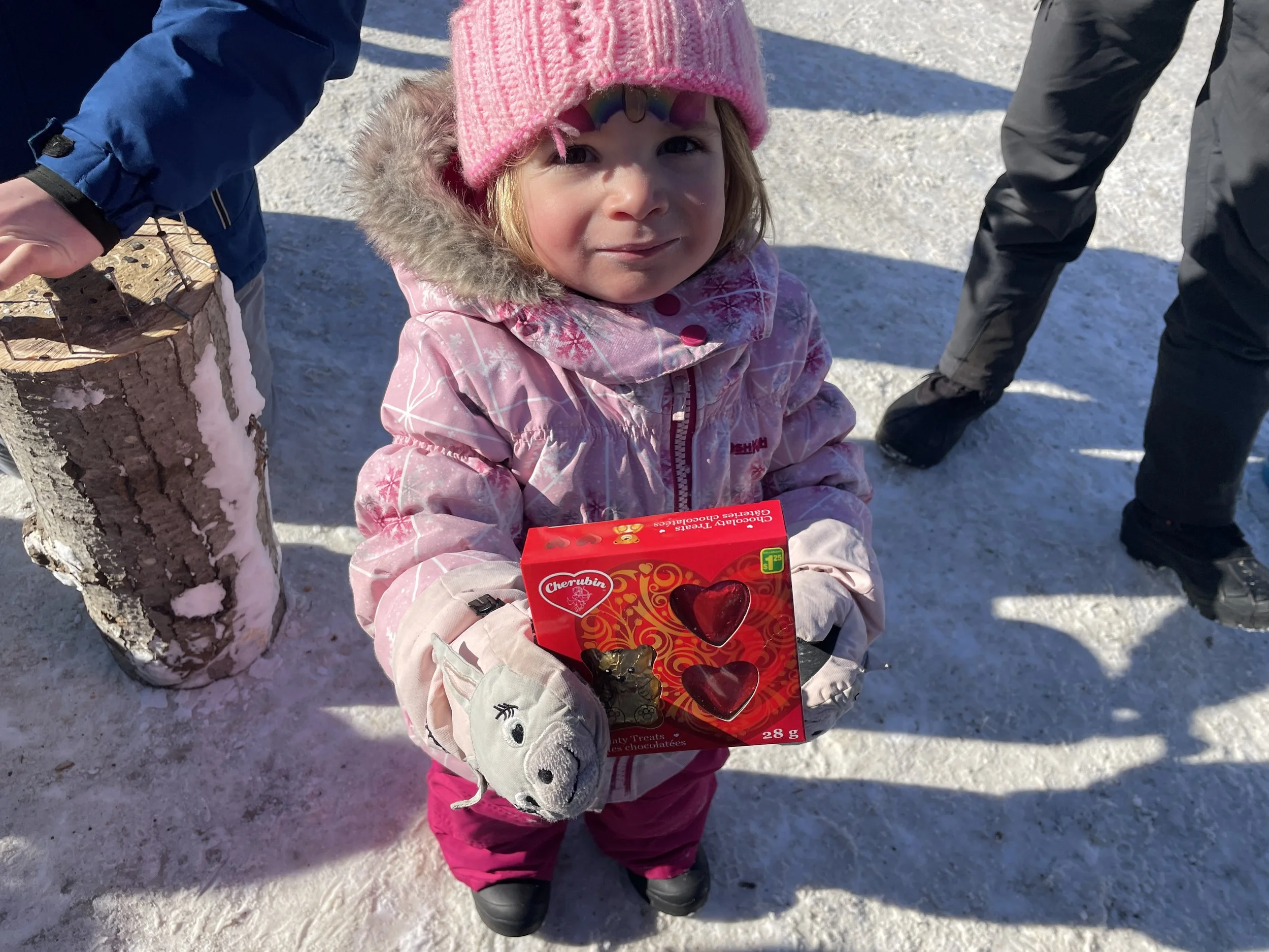 A young girl in a pink winter coat and pink knit hat holding a box of chocolate candies outdoors on snow-covered ground.