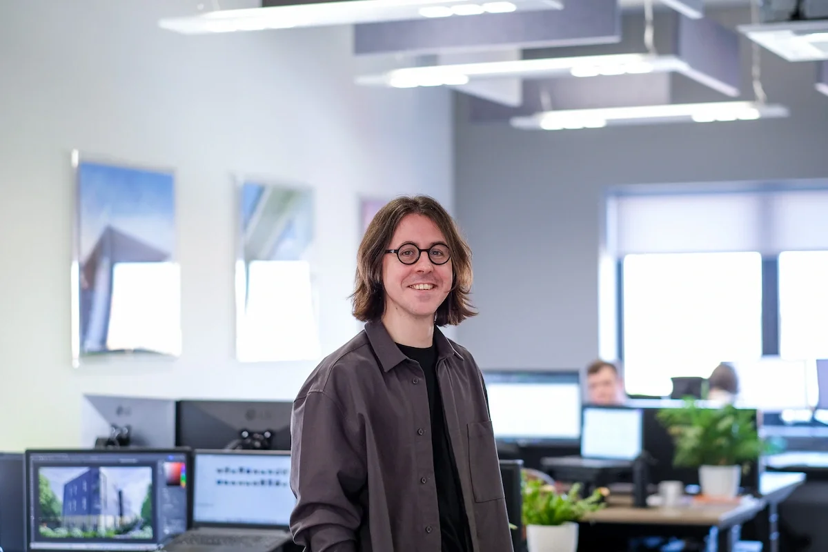 A young man with long brown hair and glasses smiling in front of an office enviroment