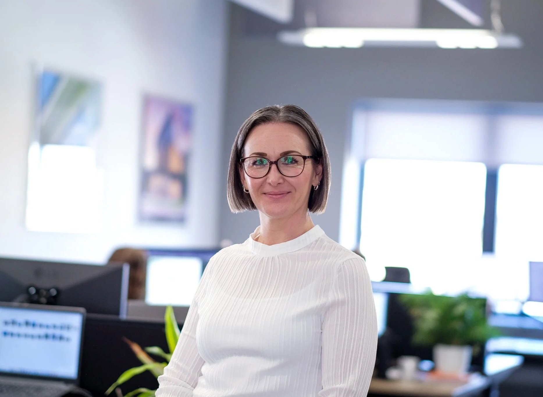 A woman wearing glasses and a white long-sleeve top standing in an office with computers and artwork in the background.