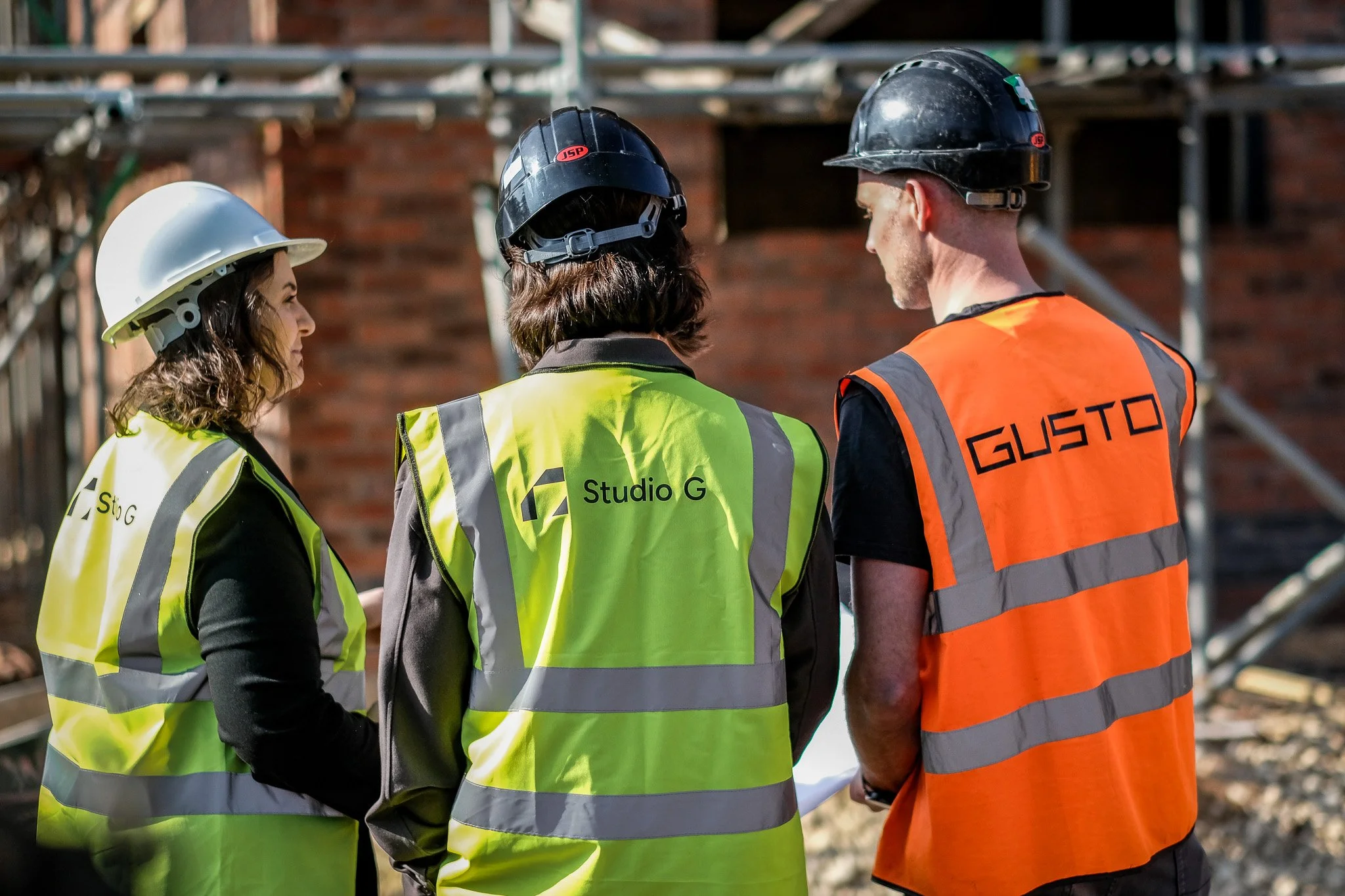 3 people, 2 of which with Studio G hi-vis vests on and the other in a GUSTO hi-vis on a construction site, looking at a building that in progress. All 3 people are wearing hard hats.