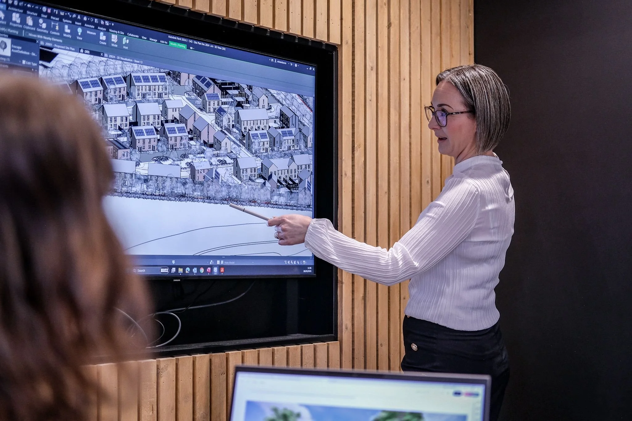 An architect in a white shirt and glasses points to a 3D housing development model on a large digital screen during a presentation