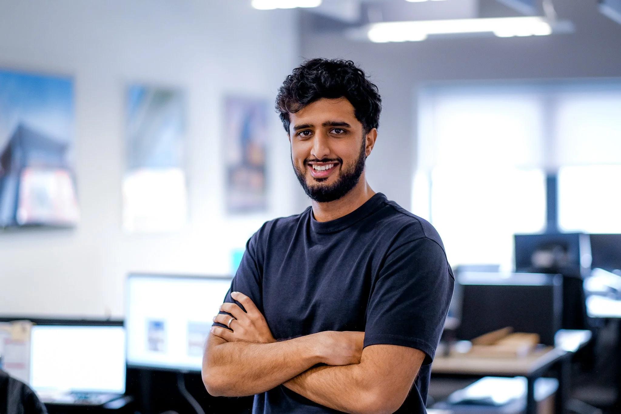 A man with dark hair and a beard smiling at the camera in an office setting, with desks, computers, and other employees in the background.