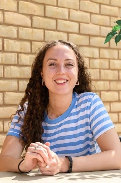 A young women with curly hair and blue striped top, smiling in front of a brick wall.