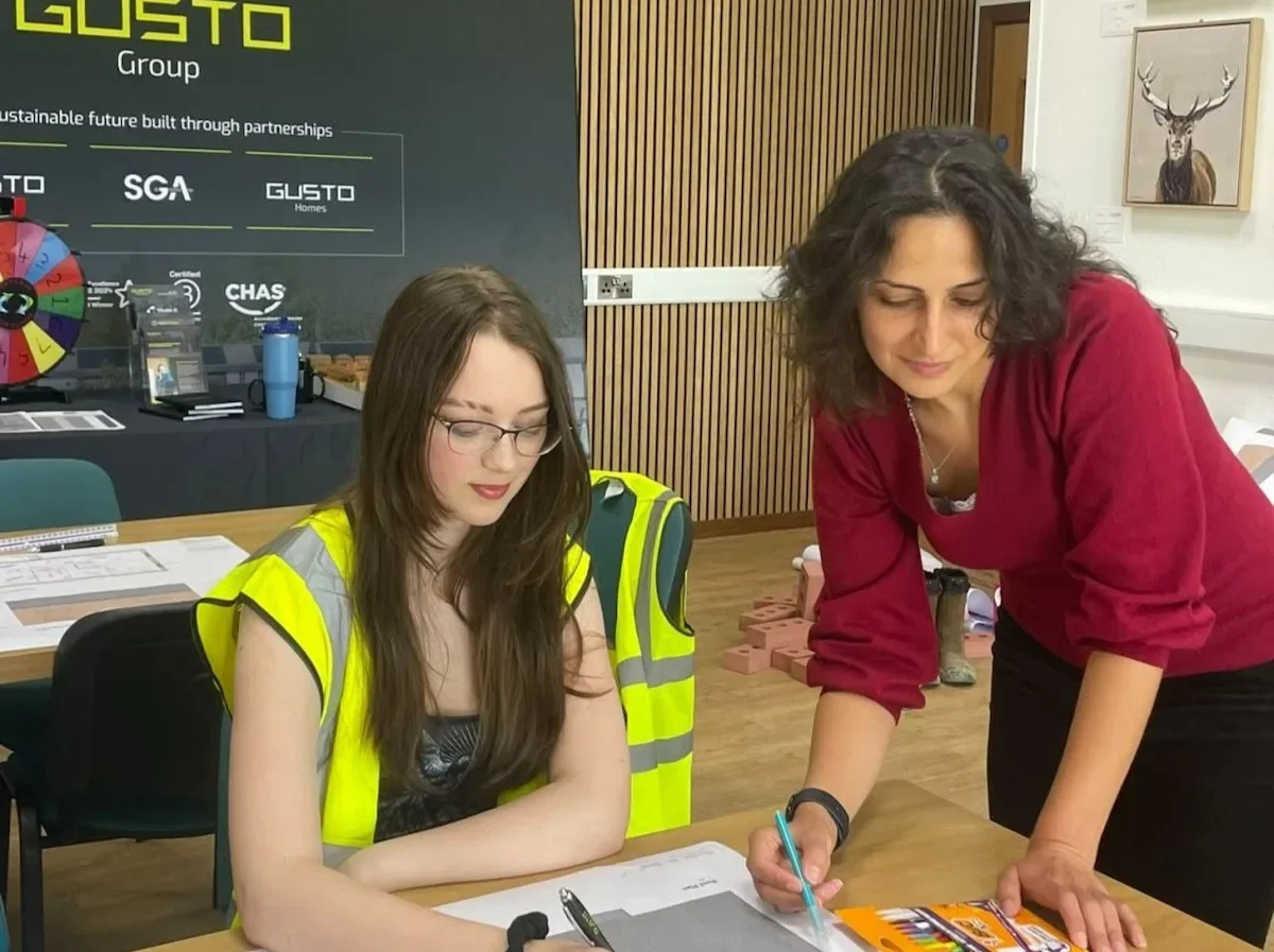 A young women in a high vis and another women leant over the table. They are both looking at a drawing.